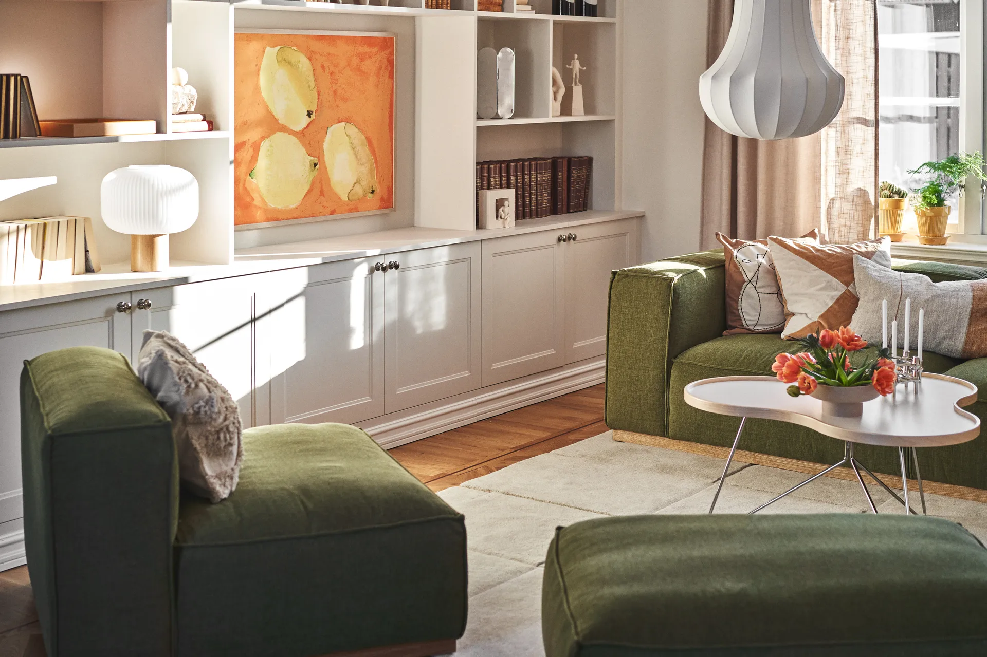 Słoneczny salon z zielonymi sofami, białym stolikiem kawowym z pomarańczowymi tulipanami i świecami, białymi półkami ze sztuką.