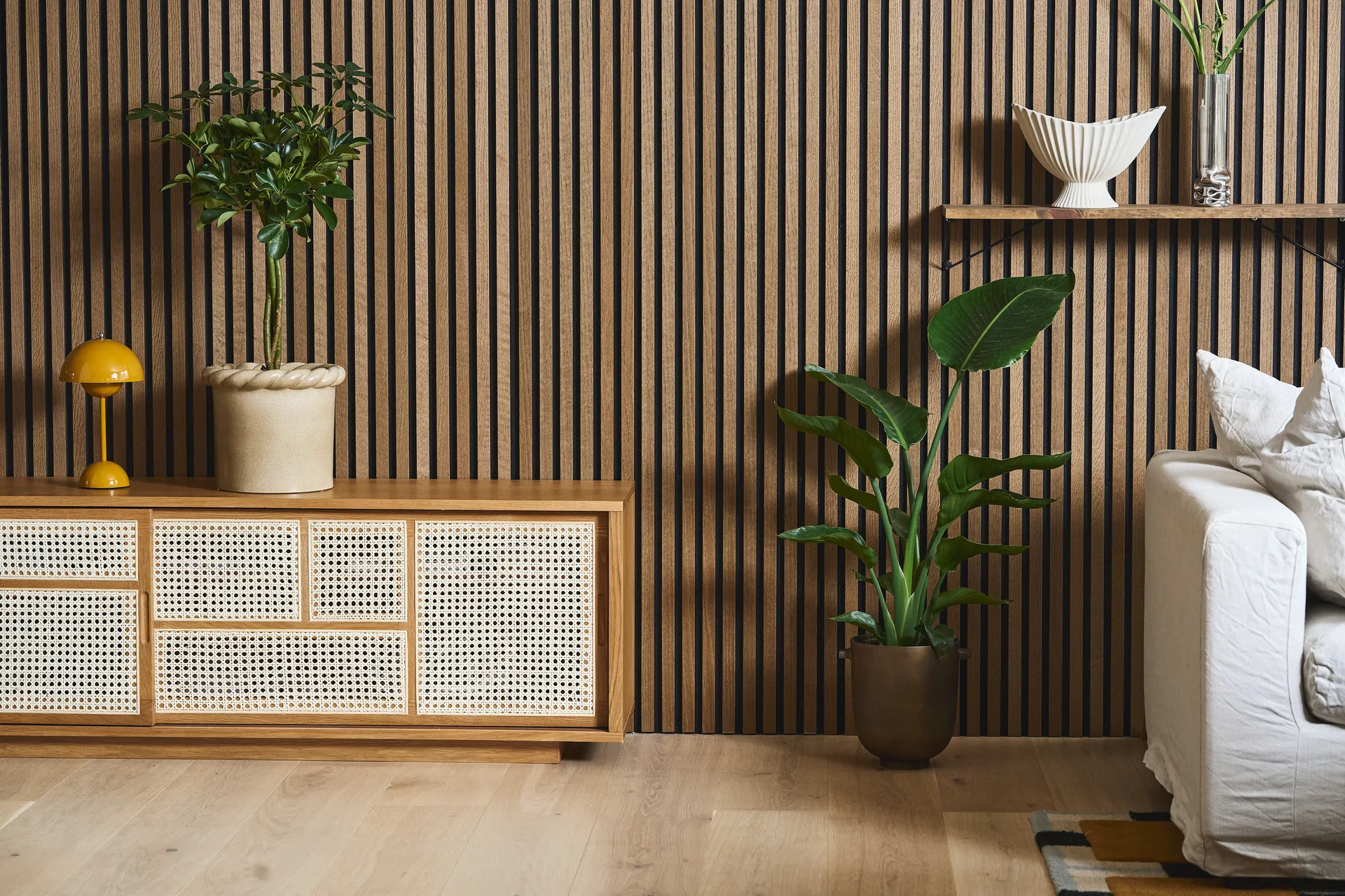 Nowoczesny salon z drewnianą ścianą lamelową, szafką rattanową, żółtą lampą, roślinami i białą sofą.