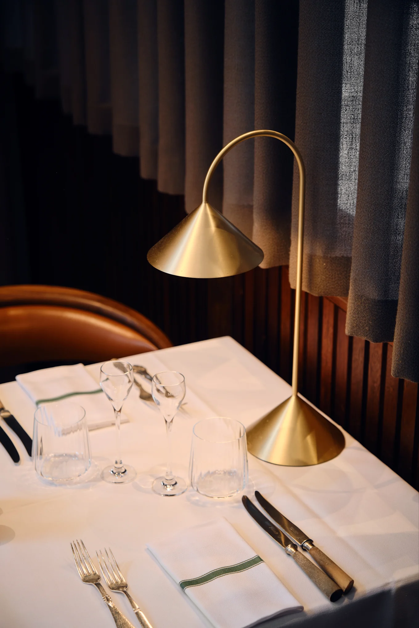 Nowoczesna złota lampa stołowa oświetla nakryty stół restauracyjny z białym obrusem, szklankami, sztućcami i serwetką z zielonym paskiem.
