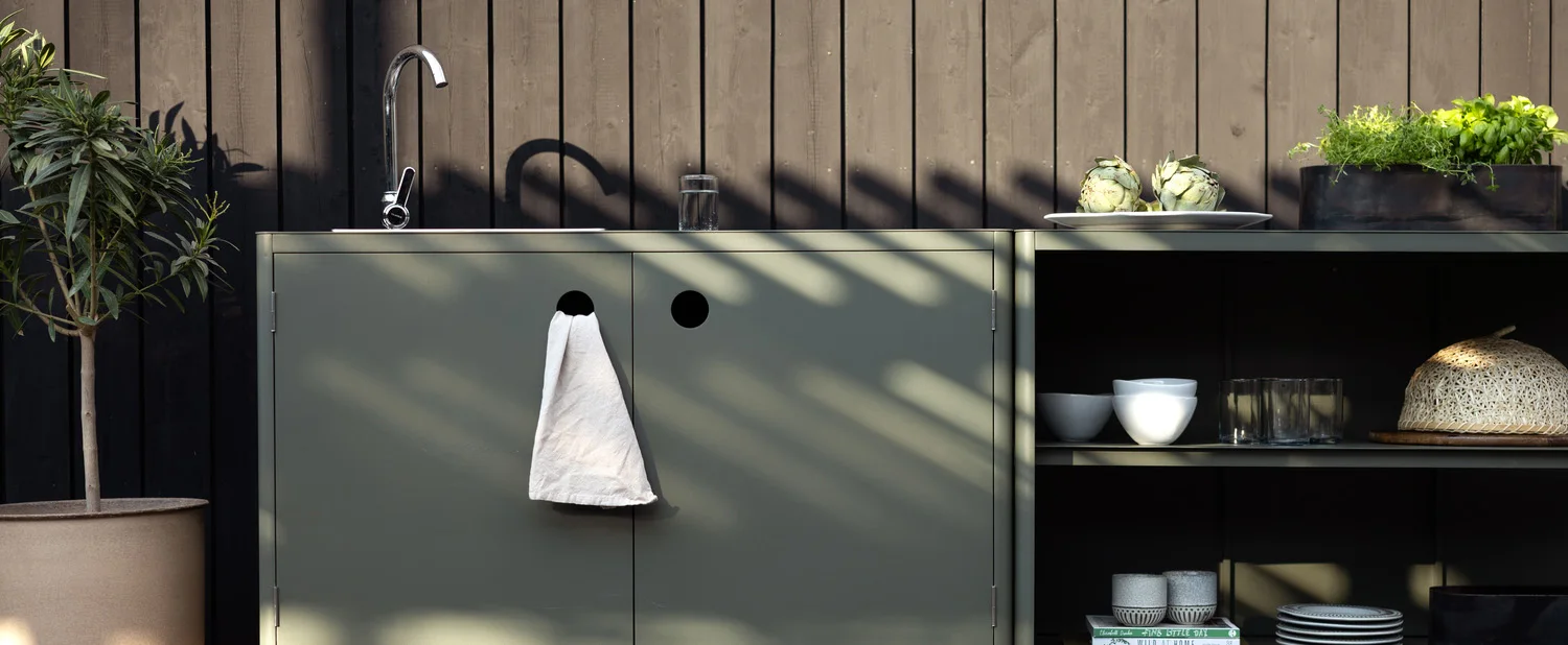 Kuchnia zewnętrzna z szałwiowo-zielonymi szafkami, zlewem, ziołami i rośliną doniczkową na tle ciemnej drewnianej ściany.