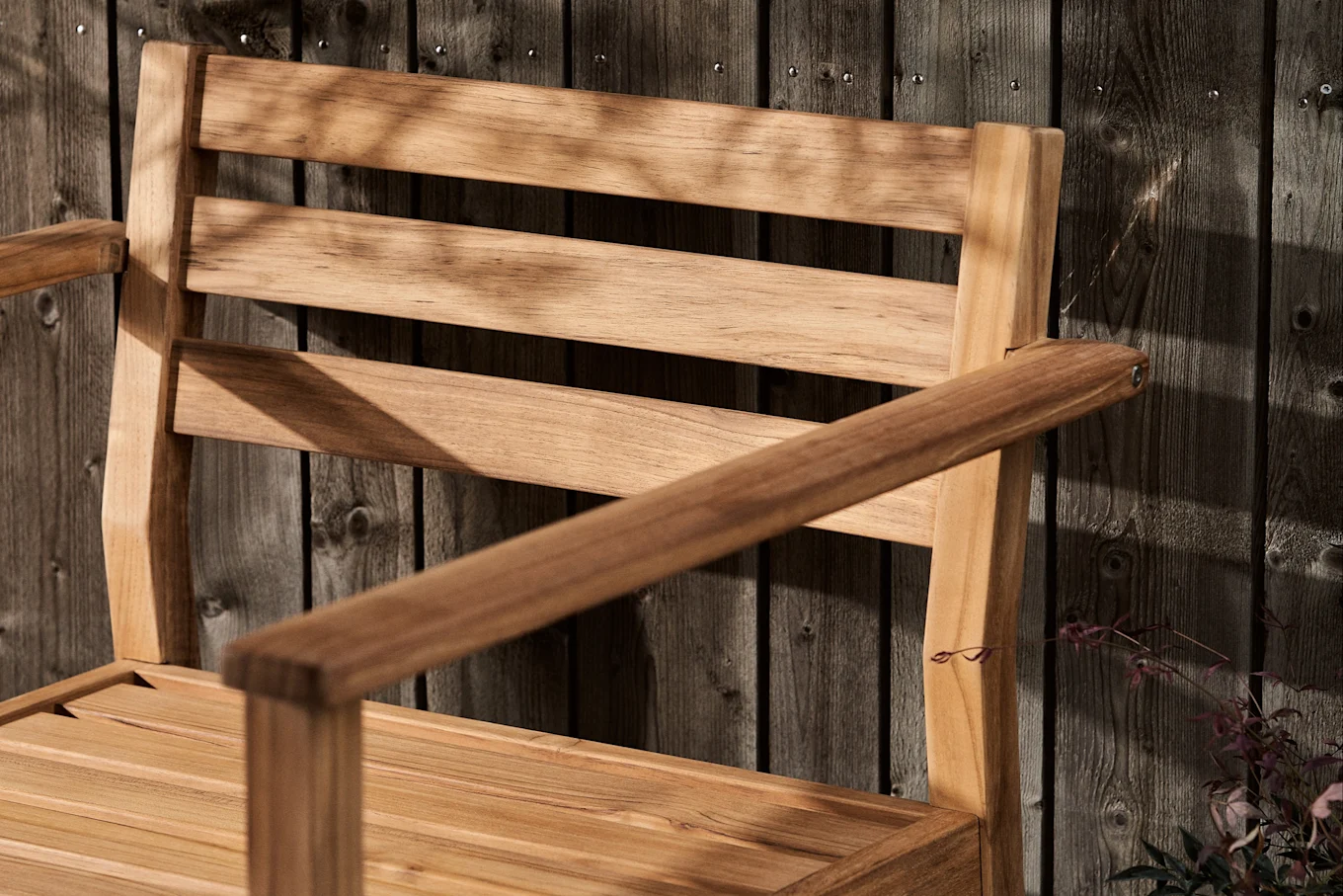 Drewniane krzesło z lamelami i podłokietnikami na tle ciemnego drewnianego płotu, pokryte cieniami liści.