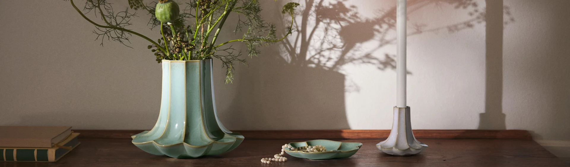 Ryflowany turkusowy wazon z liśćmi, naszyjnik z pereł w pasującym naczyniu, biała świeca w ryflowanym świeczniku i książki na drewnianej powierzchni.