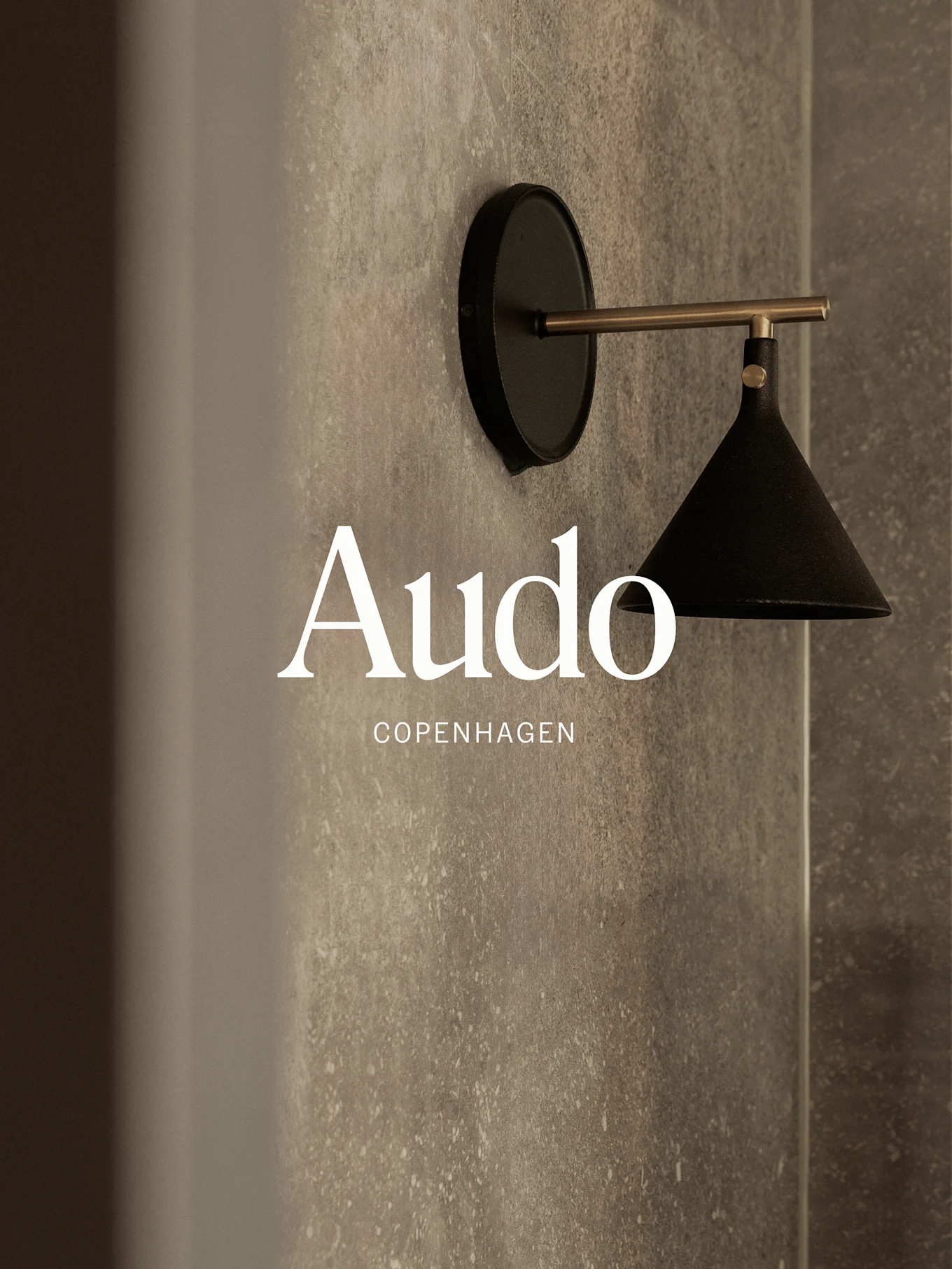 Czarna lampa ścienna z mosiężnym ramieniem na tle teksturowanej szarej ściany, z napisem 'Audo Copenhagen'.