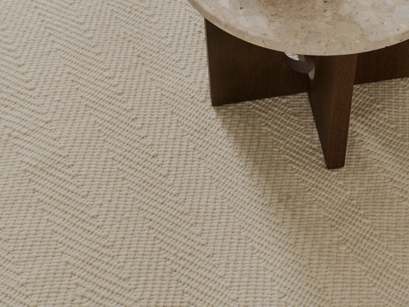 Zbliżenie na kremowy, teksturowany dywan z wzorem w jodełkę, obok stołu z kamiennym blatem.