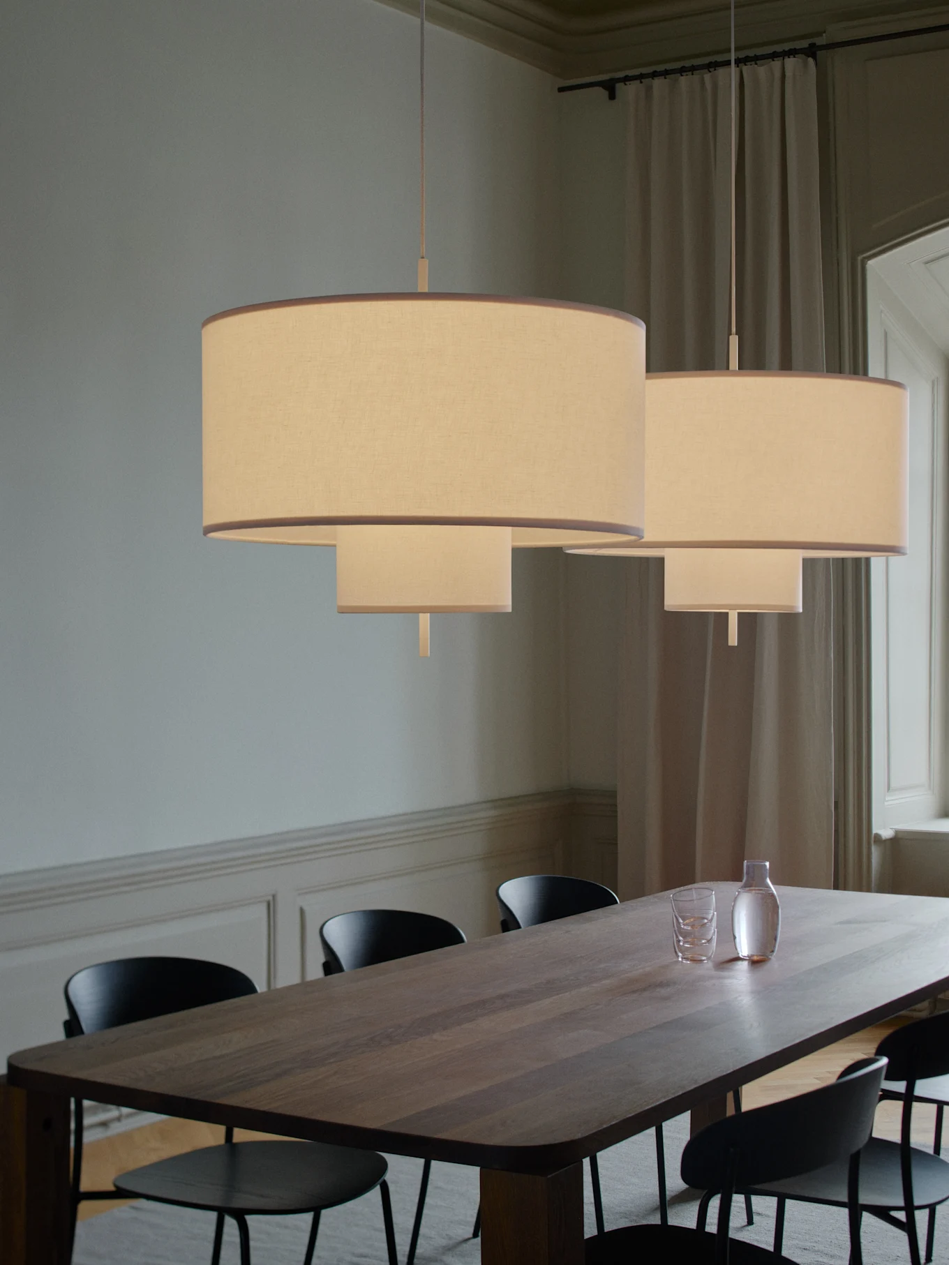 Dwie piętrowe lampy wiszące z tkaniny oświetlają długi drewniany stół jadalny z czarnymi krzesłami, karafką i szklankami.
