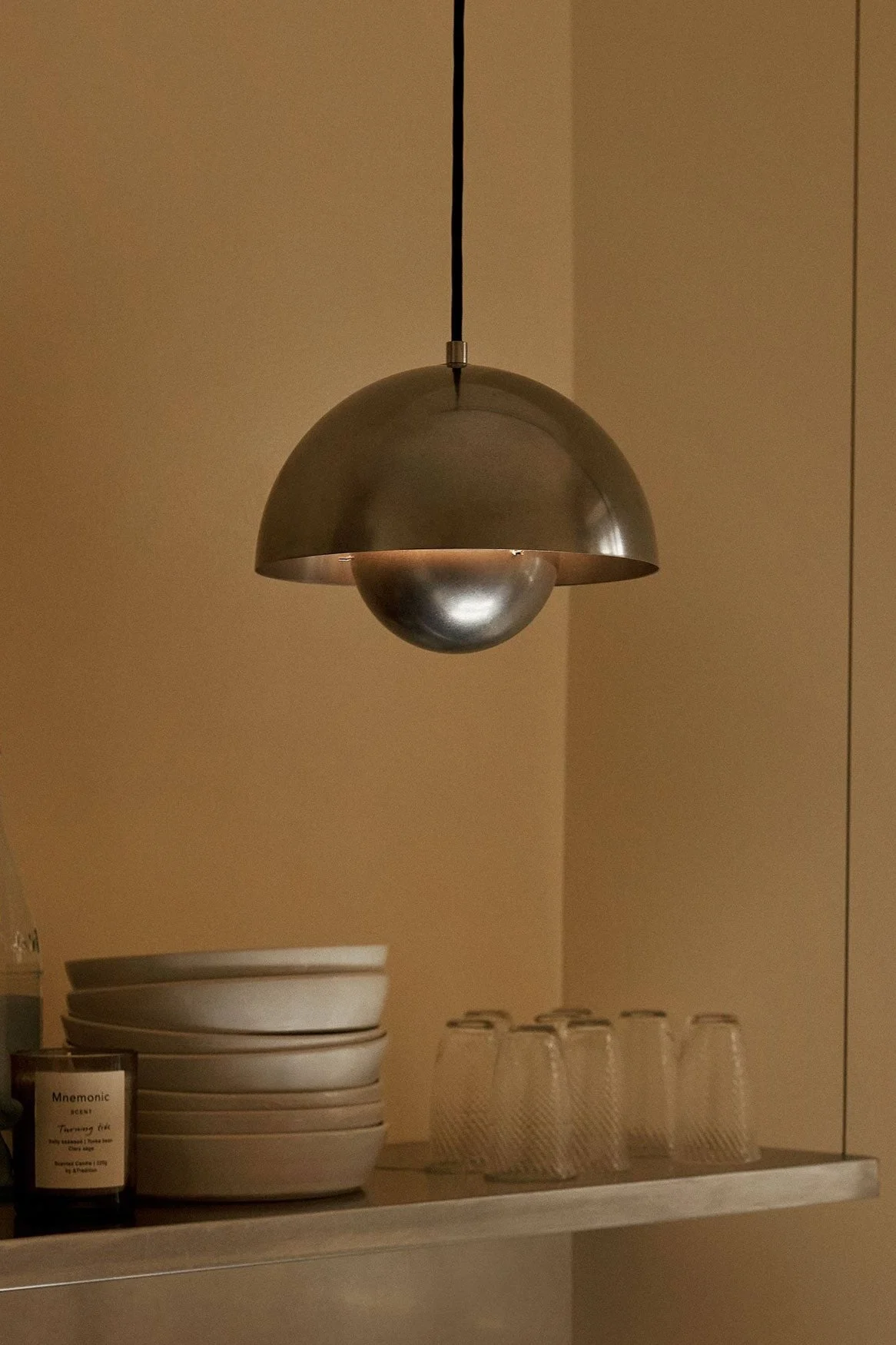Kopułowa metalowa lampa wisząca wisi nad półką z miskami, szklankami, butelkami i świecą na tle ciepłej brązowej ściany.