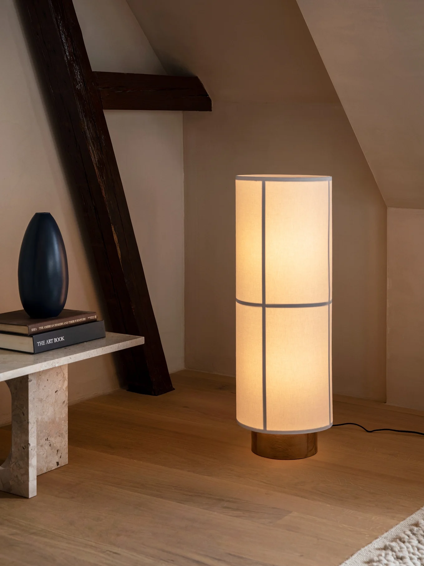 Wysoka cylindryczna lampa podłogowa z jasnym kloszem i drewnianą podstawą oświetla pokój z ławką i belką.