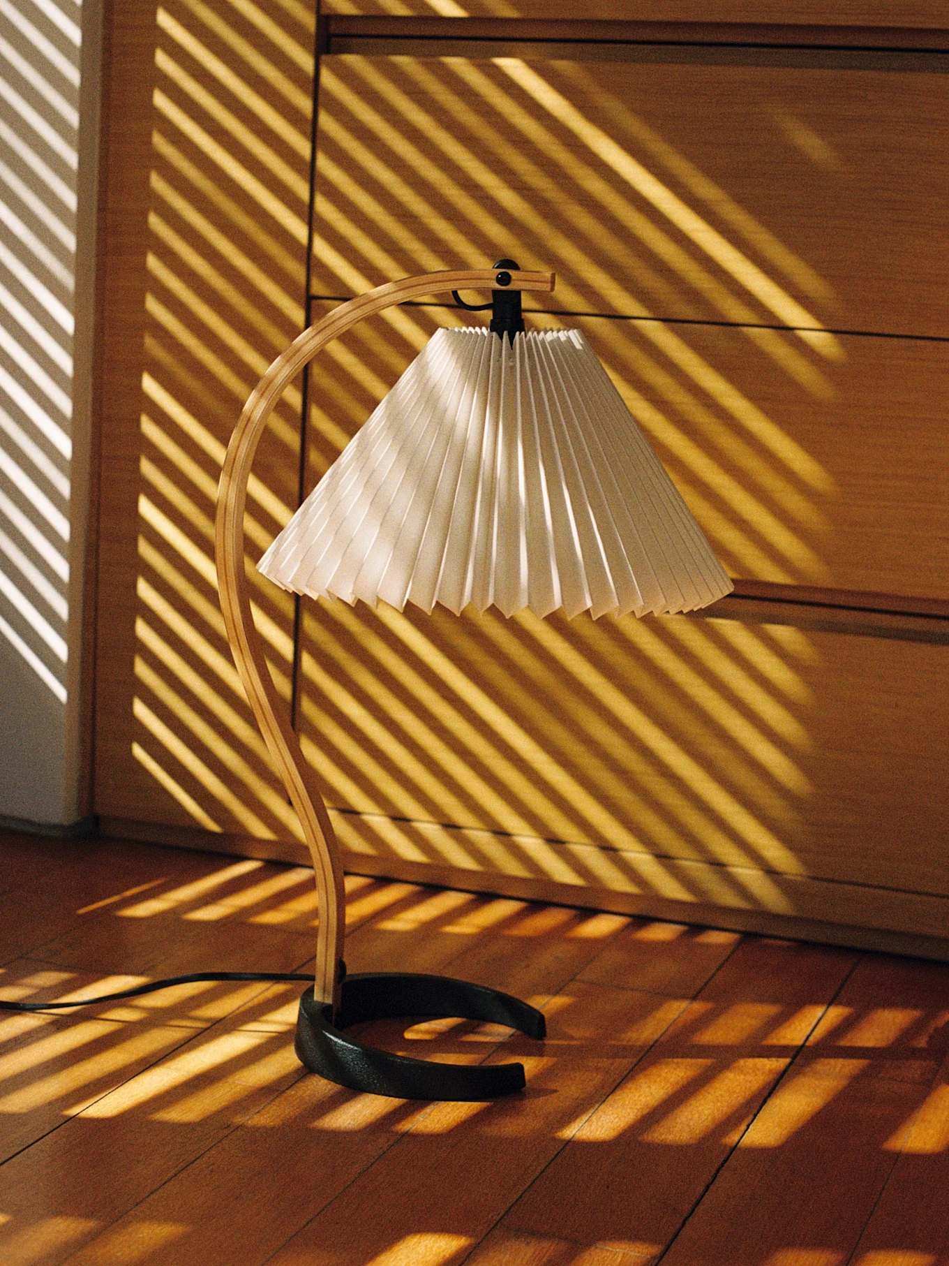 Lampa biurkowa z drewnianą podstawą i plisowanym kloszem, oświetlona ukośnymi promieniami słońca na drewnianej podłodze.