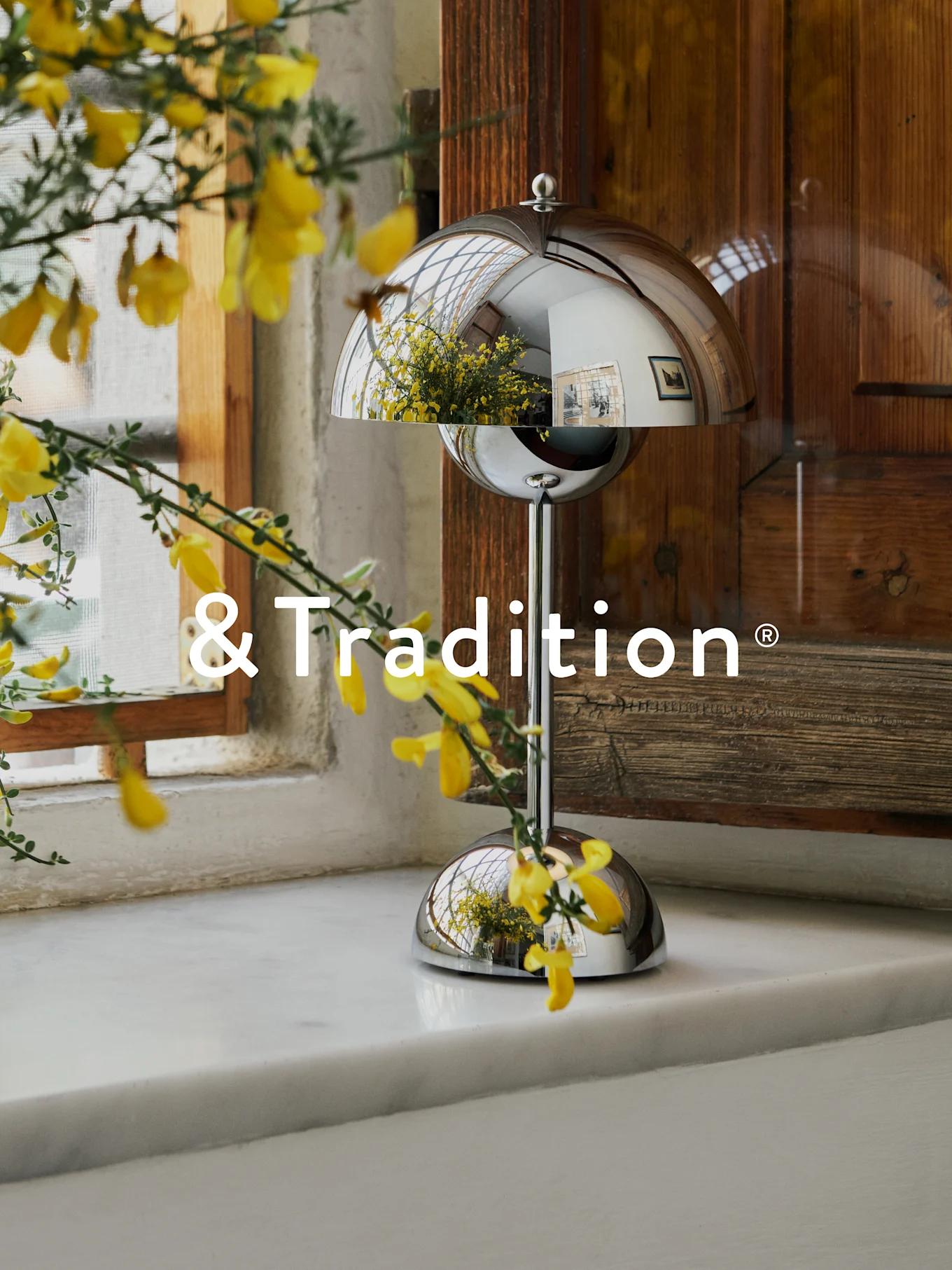 Srebrna, kopułkowa lampa stołowa na białym parapecie, z żółtymi kwiatami na pierwszym planie. Lampa odbija okno i kwiaty. Logo '&Tradition'.
