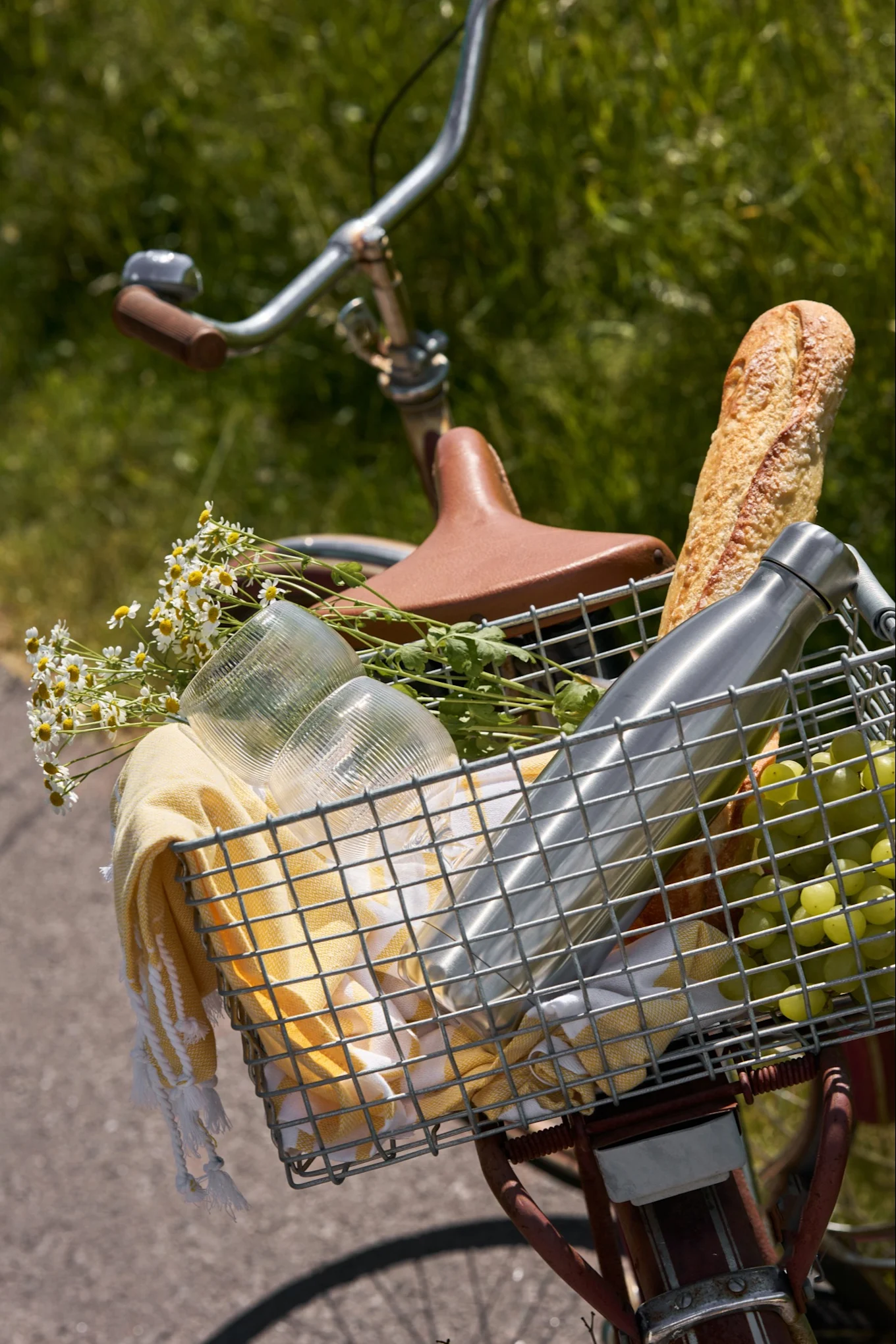 Koszyk rowerowy wypełniony artykułami piknikowymi: bagietka, butelka wody, winogrona, szklanki, żółty ręcznik i białe stokrotki.