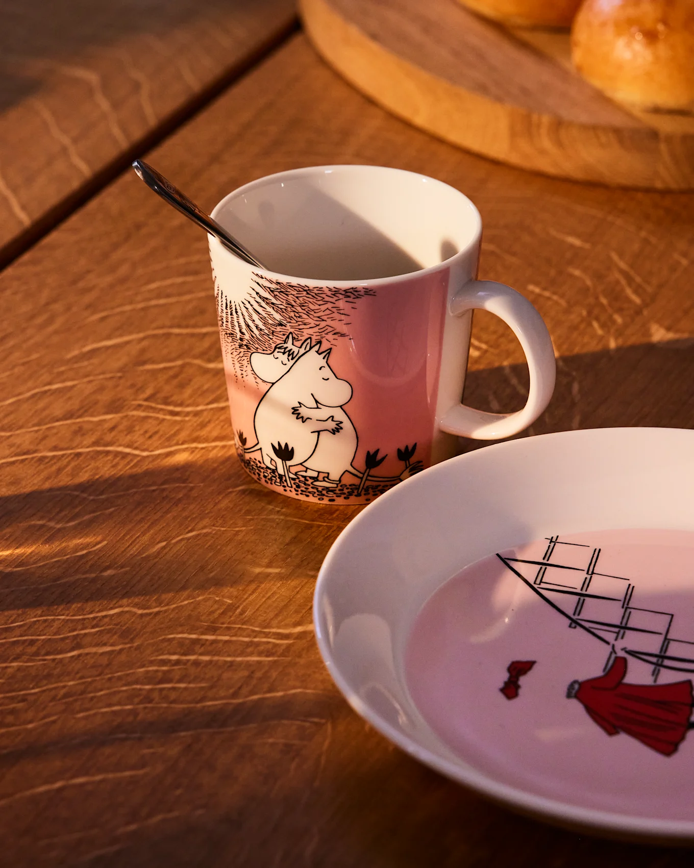 Różowy kubek Moomin z Muminkiem i Migotką, obok pasującego talerza z żaglowcem, na drewnianym stole.