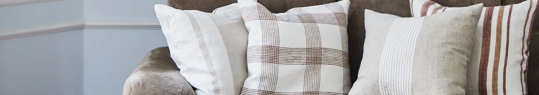 Zbliżenie trzech poduszek na brązowej sofie, z białymi, beżowymi i kraciastymi wzorami w brązowe i czerwone pasy.