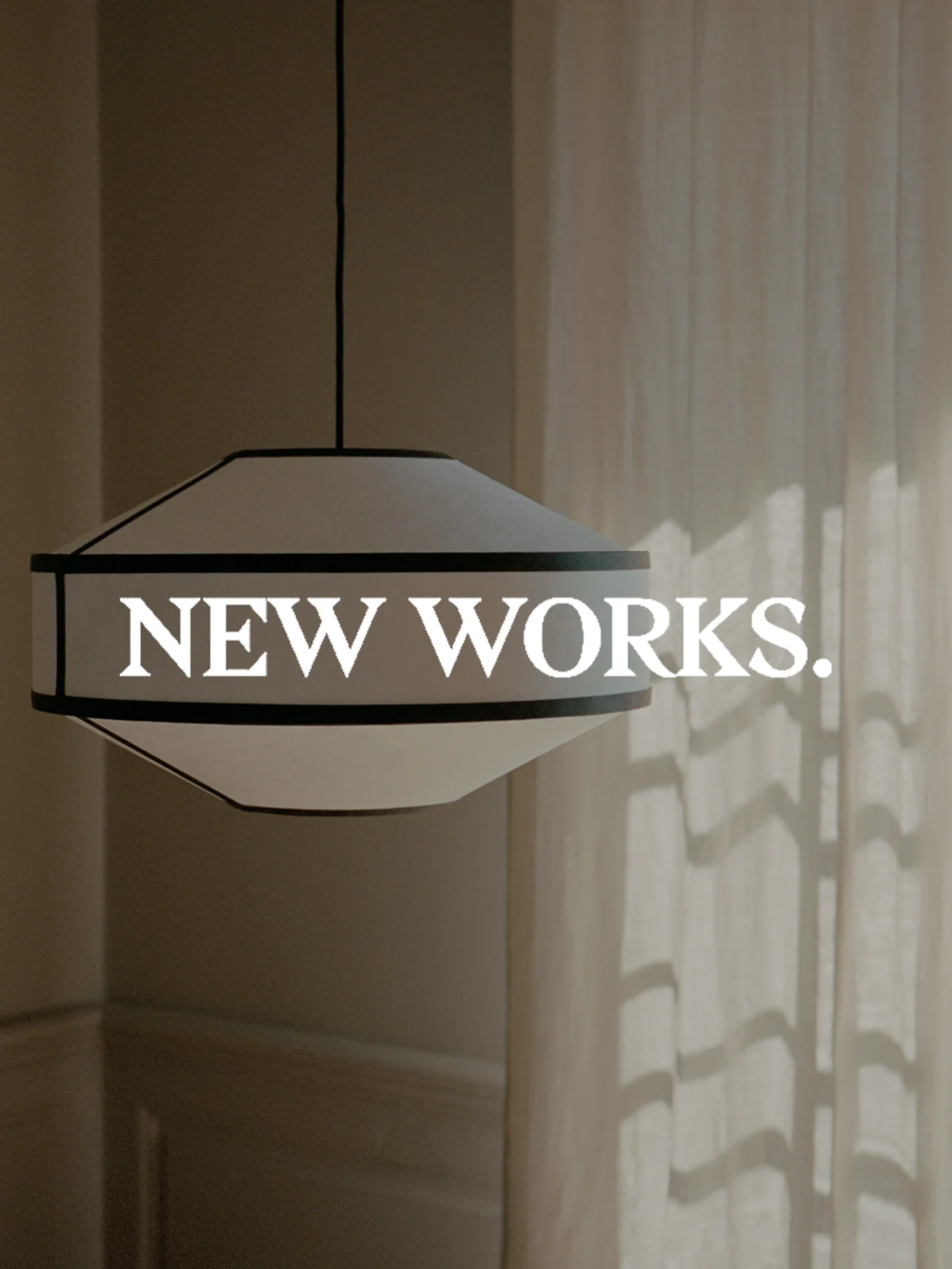 Szeroka, biała lampa wisząca z ciemnym obrzeżeniem, z napisem "NEW WORKS.". Słońce rzuca cienie na zasłonę.