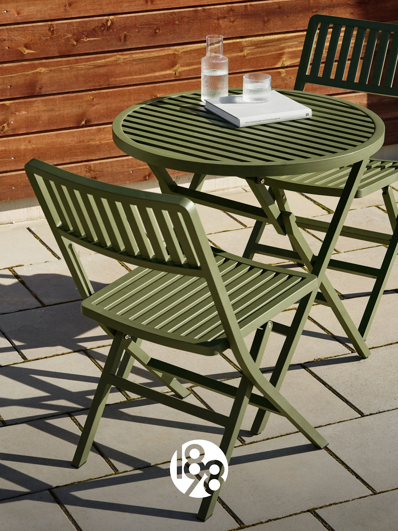 Dwa zielone składane krzesła z listewkami i pasujący stół na patio z książką i szklankami wody.