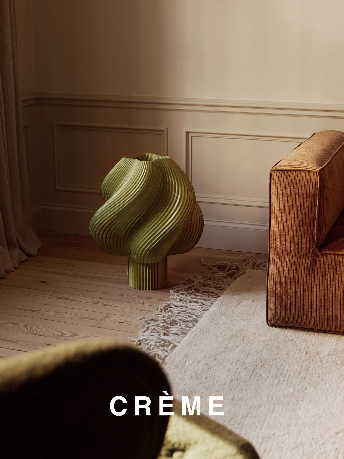 Jasnozielona, skręcona żebrowana lampa na drewnianej podłodze, obok brązowej żebrowanej sofy i beżowego dywanu shaggy.