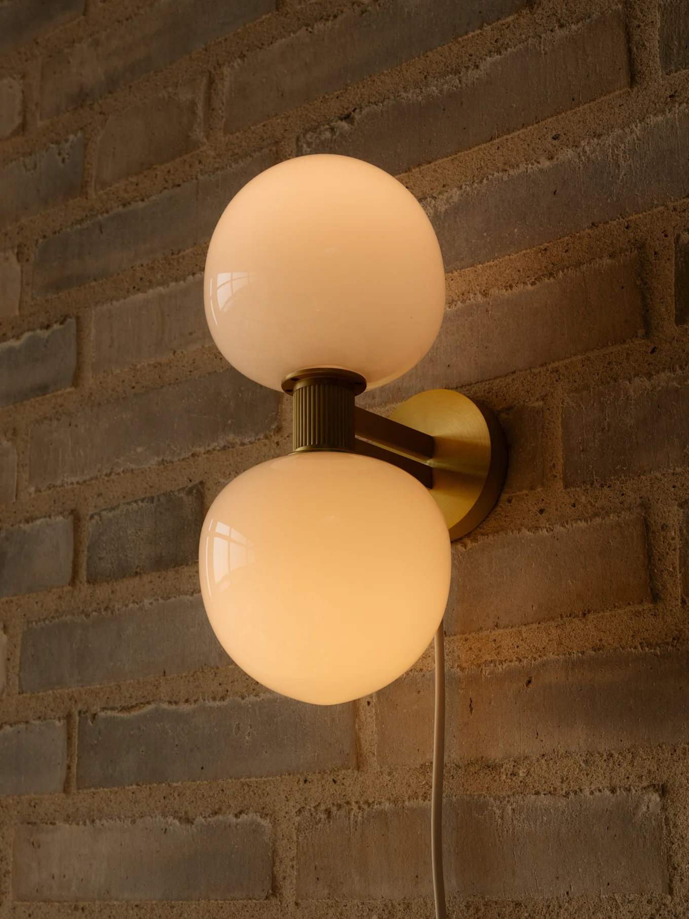Lampa ścienna z dwoma mlecznobiałymi kulistymi kloszami i mosiężnym mocowaniem na ceglanej ścianie.