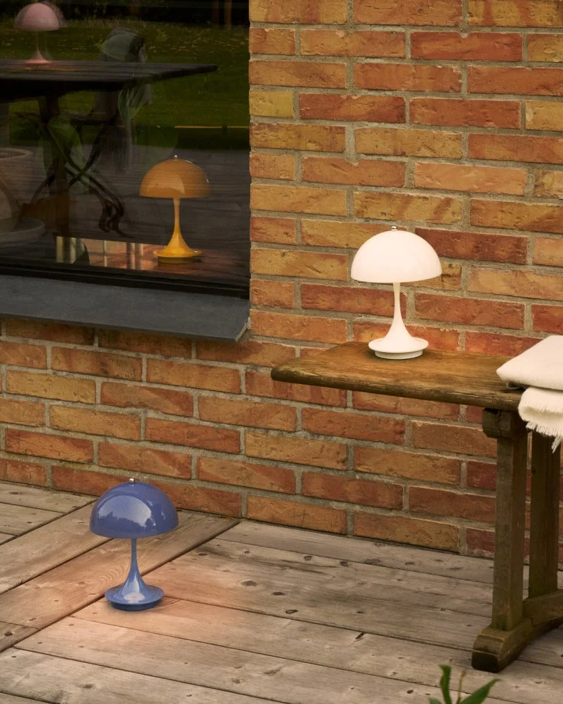 Trzy lampy w kształcie grzyba w kolorze białym, niebieskim i żółtym na drewnianym patio przy ceglanej ścianie i oknie.