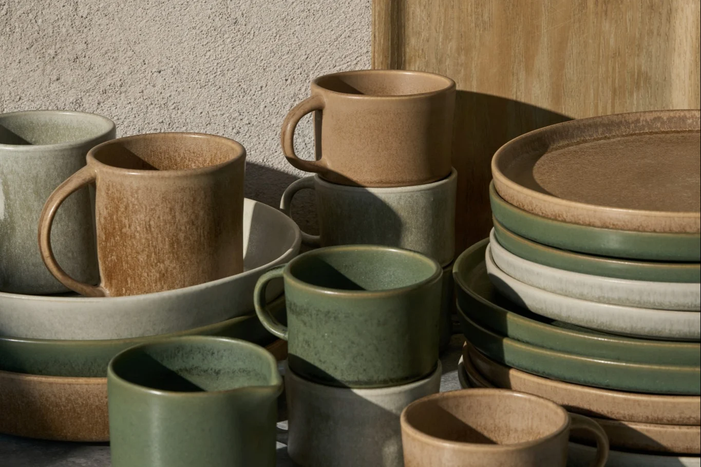 Ułożone ceramiczne kubki, miski i talerze w kolorze beżowym, jasnoszarym i szałwiowym zielonym, z rustykalnym wykończeniem.