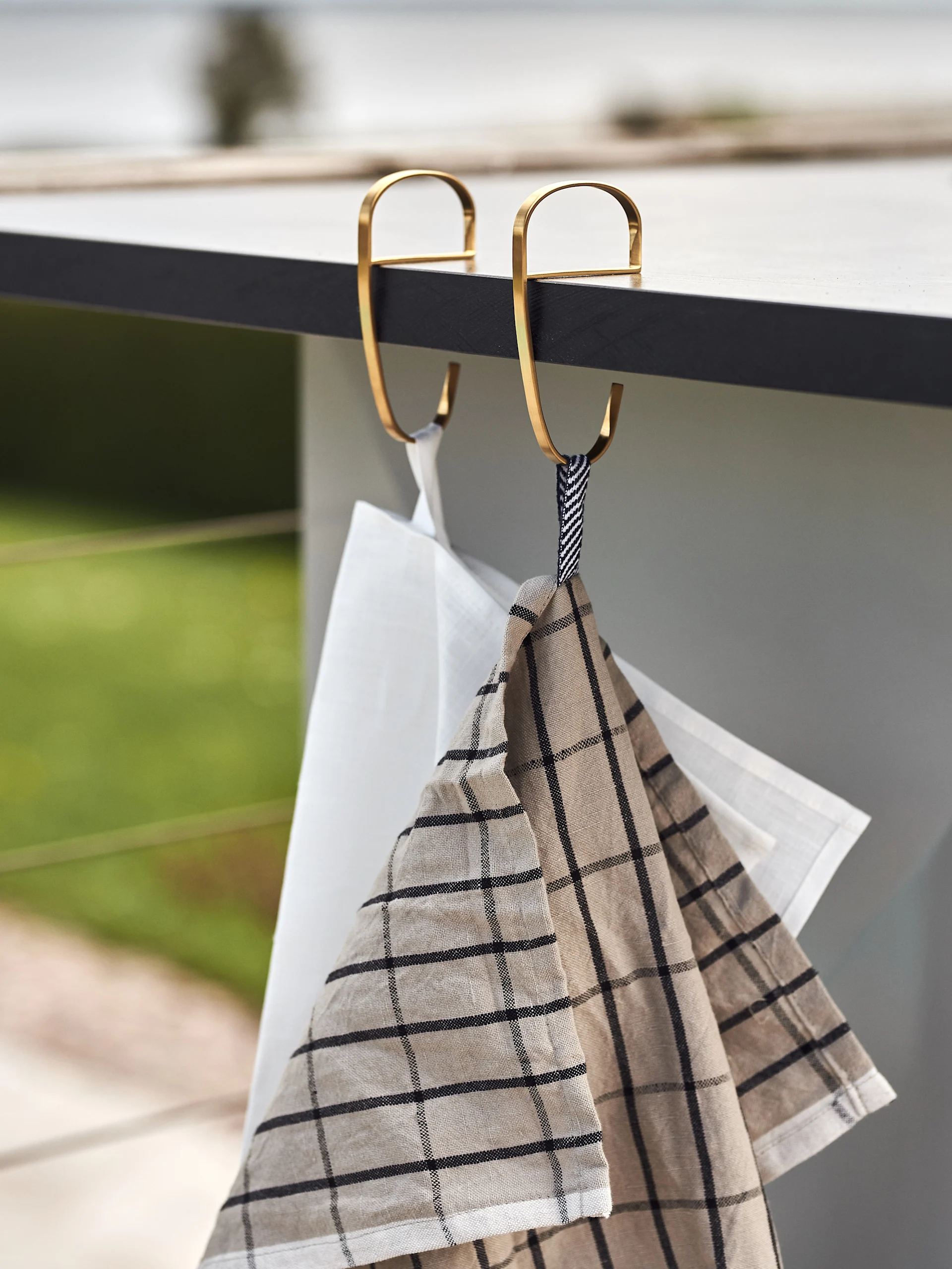 Dwa ręczniki kuchenne, jeden biały i jeden beżowy w kratę, wiszą na złotych haczykach S na ciemnym blacie.