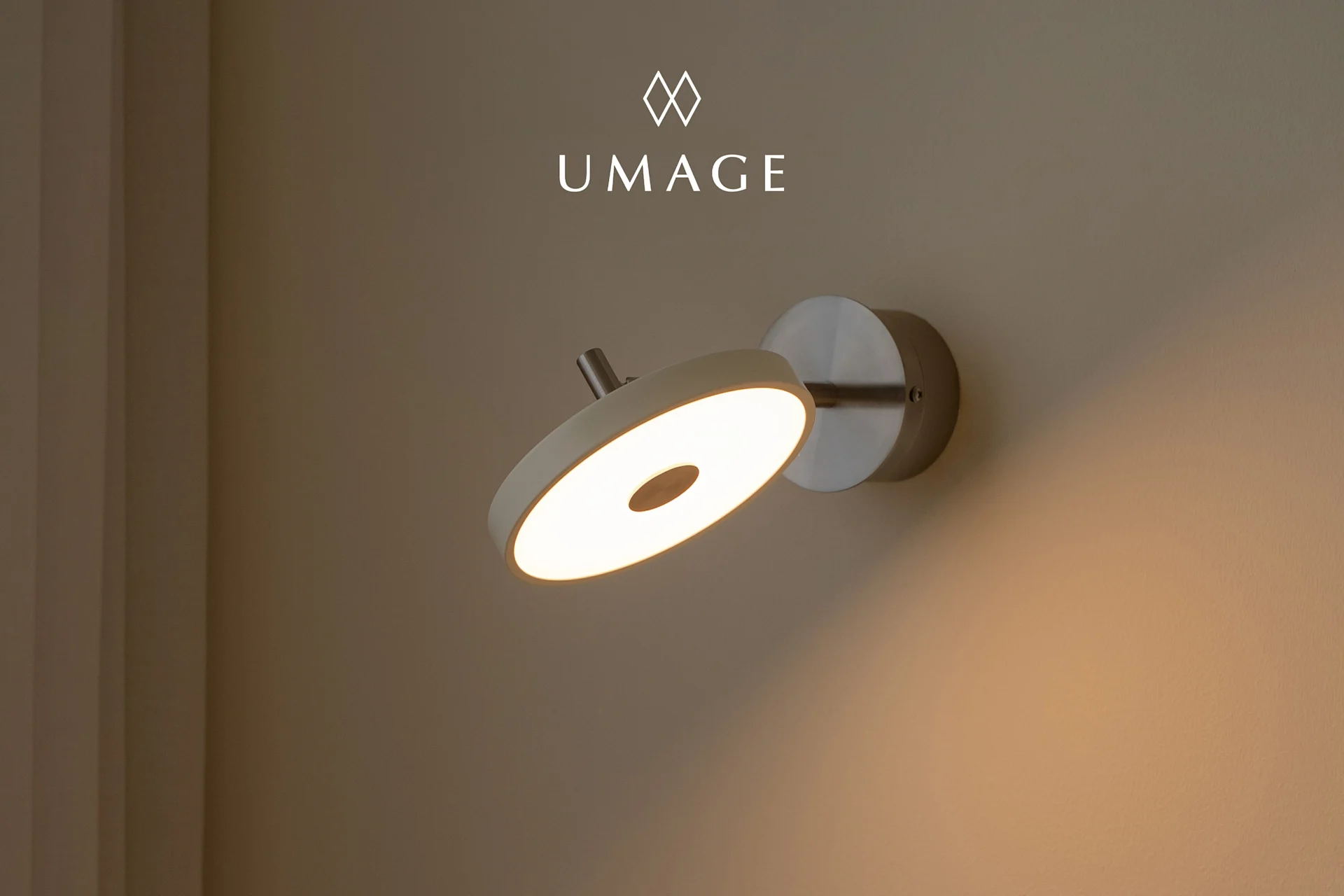 Nowoczesna okrągła lampa ścienna z ciepłym, świecącym światłem, zamontowana na jasnobrązowej ścianie. Logo UMA u góry.