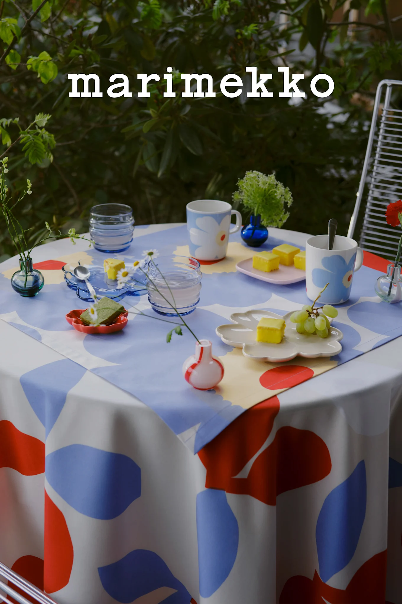 Logo Marimekko. Stół na zewnątrz z obrusem w niebieskie, czerwone i białe abstrakcyjne wzory, kwiatowymi kubkami, żółtymi ciastami, winogronami i małymi wazonami.
