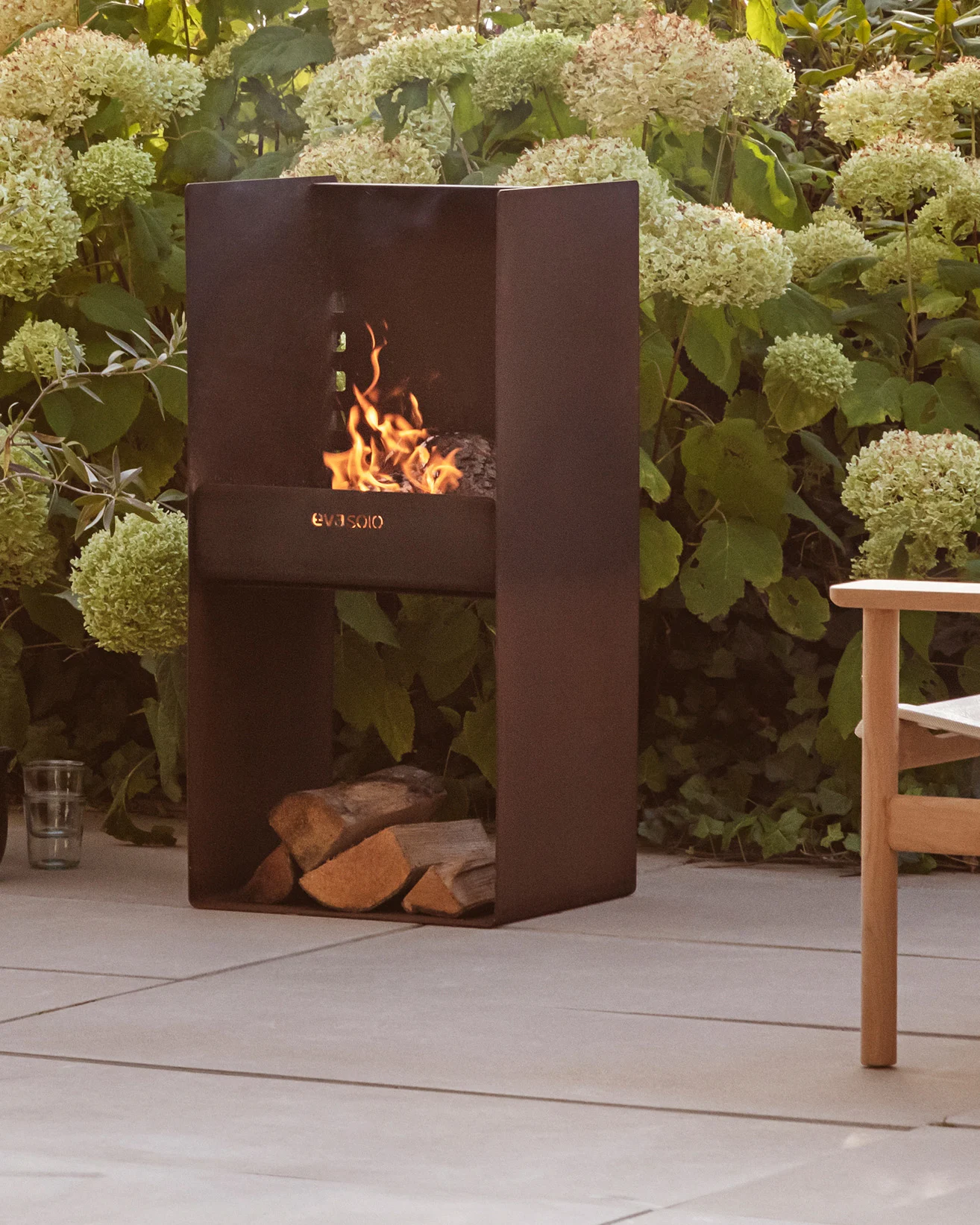 Nowoczesny ciemny metalowy kominek zewnętrzny z płonącym ogniem, ułożonymi pod nim kłodami, otoczony zielonymi roślinami i patio.