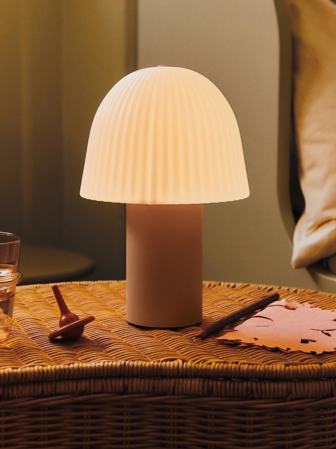 Świecąca lampa w kształcie grzyba z plisowanym kloszem na wiklinowym stoliku nocnym z wodą, bączkiem i papierem.