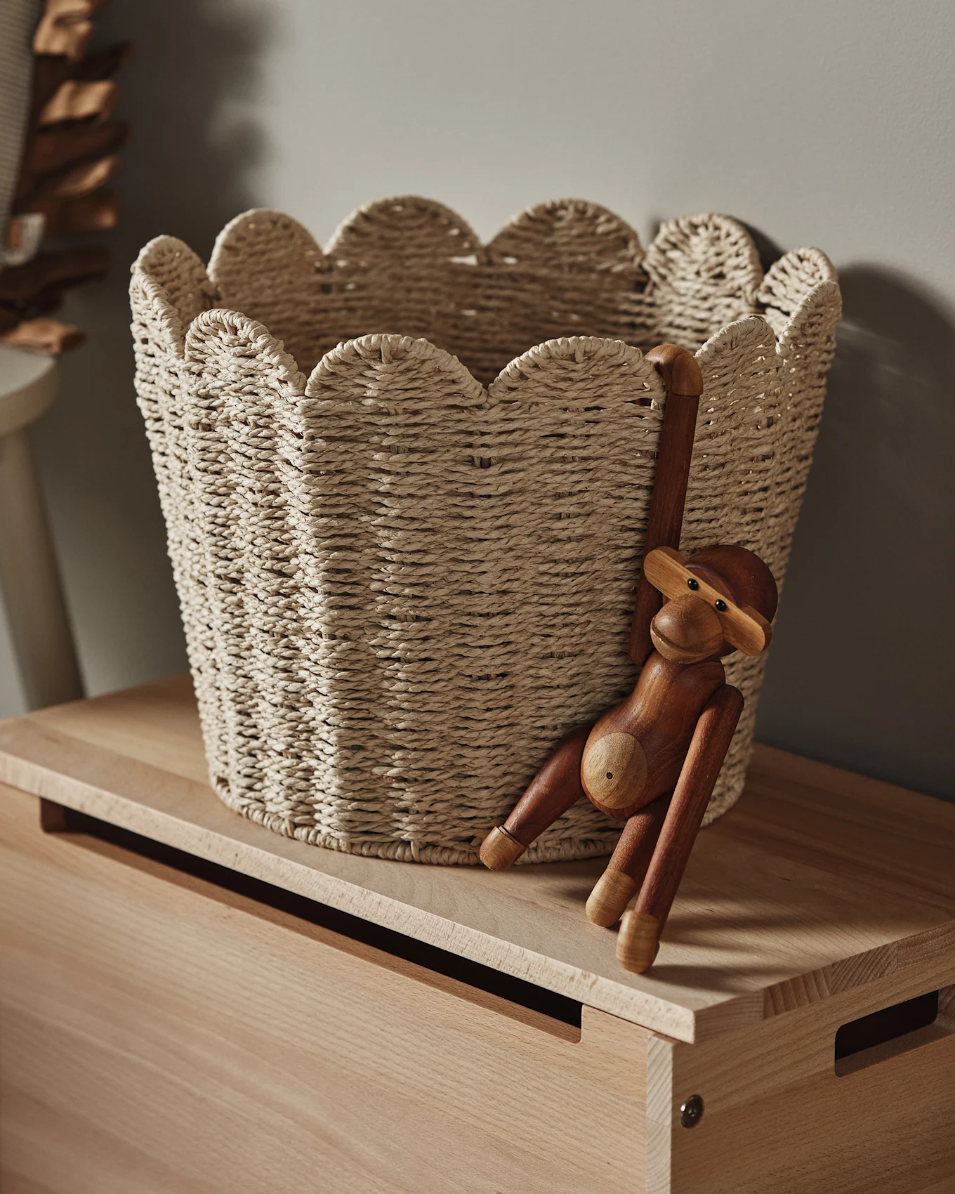 Jasny pleciony kosz z falistym brzegiem stoi na drewnianej powierzchni, z drewnianą figurką małpy zwisającą z boku.