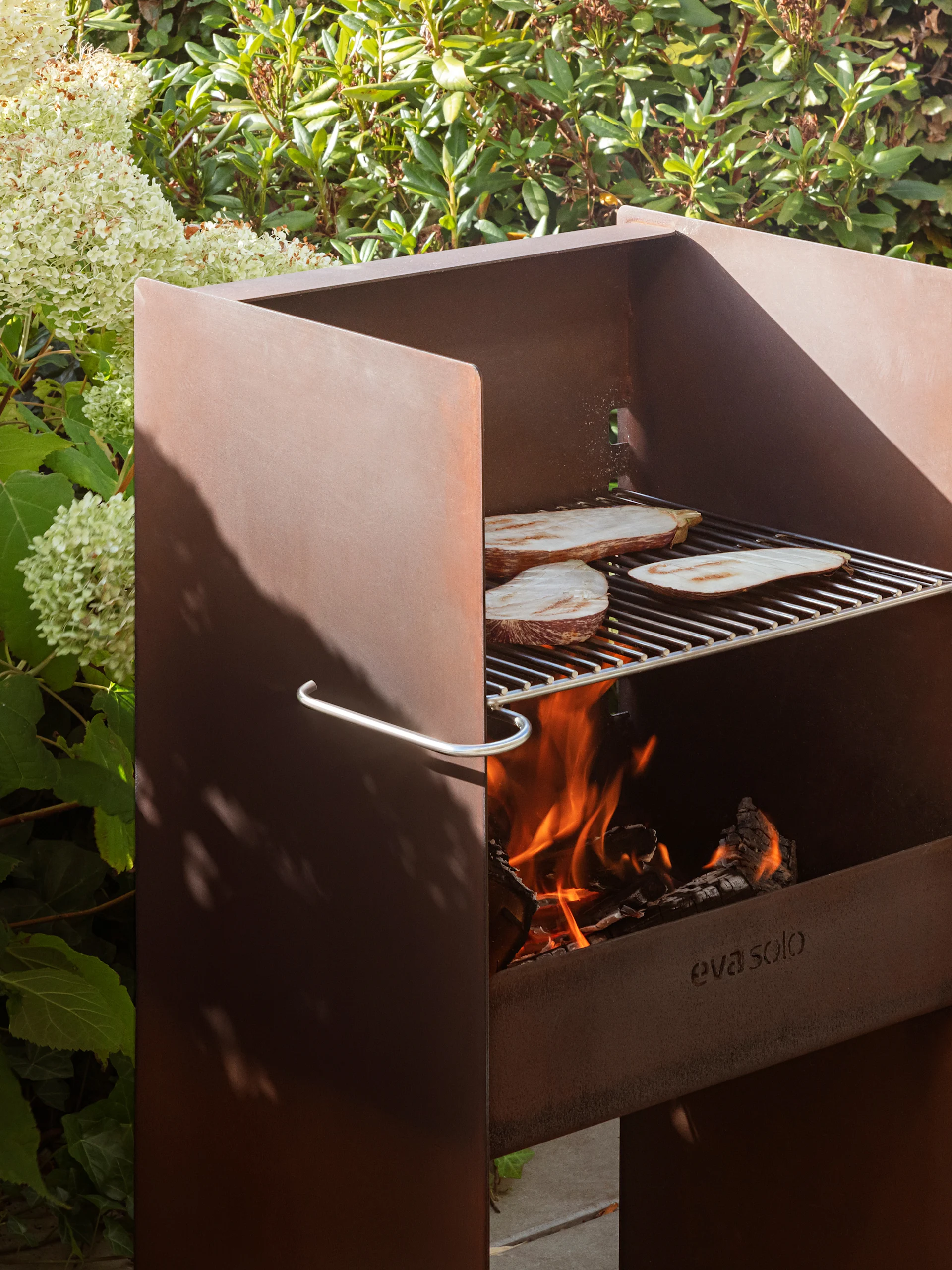 Zardzewiały metalowy grill ogrodowy Eva Solo z bakłażanem na ruszcie, płonącym ogniem i bujnym listowiem.