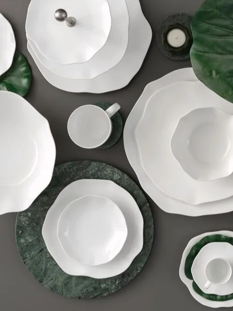Kolekcja białej zastawy ceramicznej z falistymi brzegami, ułożona z zielonymi liśćmi i marmurowymi podkładkami na ciemnej powierzchni.