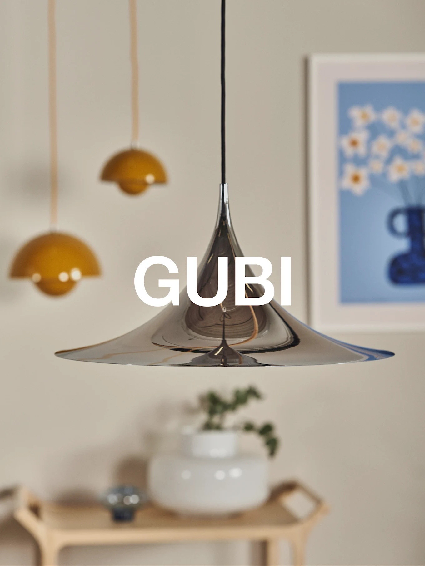Polerowana chromowana lampa wisząca stożkowa z logo GUBI, na rozmytym tle z innymi lampami i sztuką.
