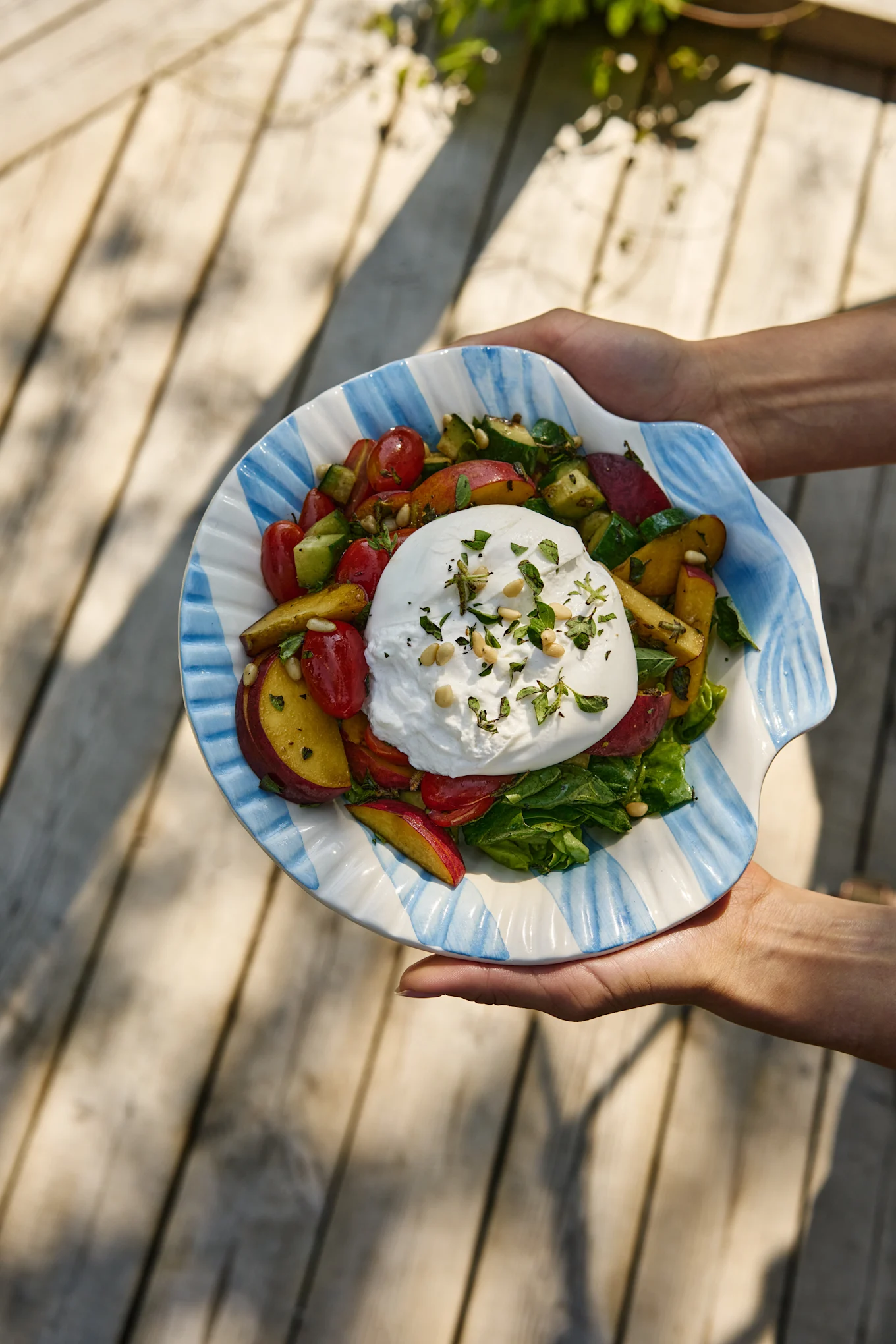 Dłonie trzymają sałatkę burrata z pomidorami, brzoskwiniami, ogórkiem i zieleniną w paskowanej muszlowej misce.