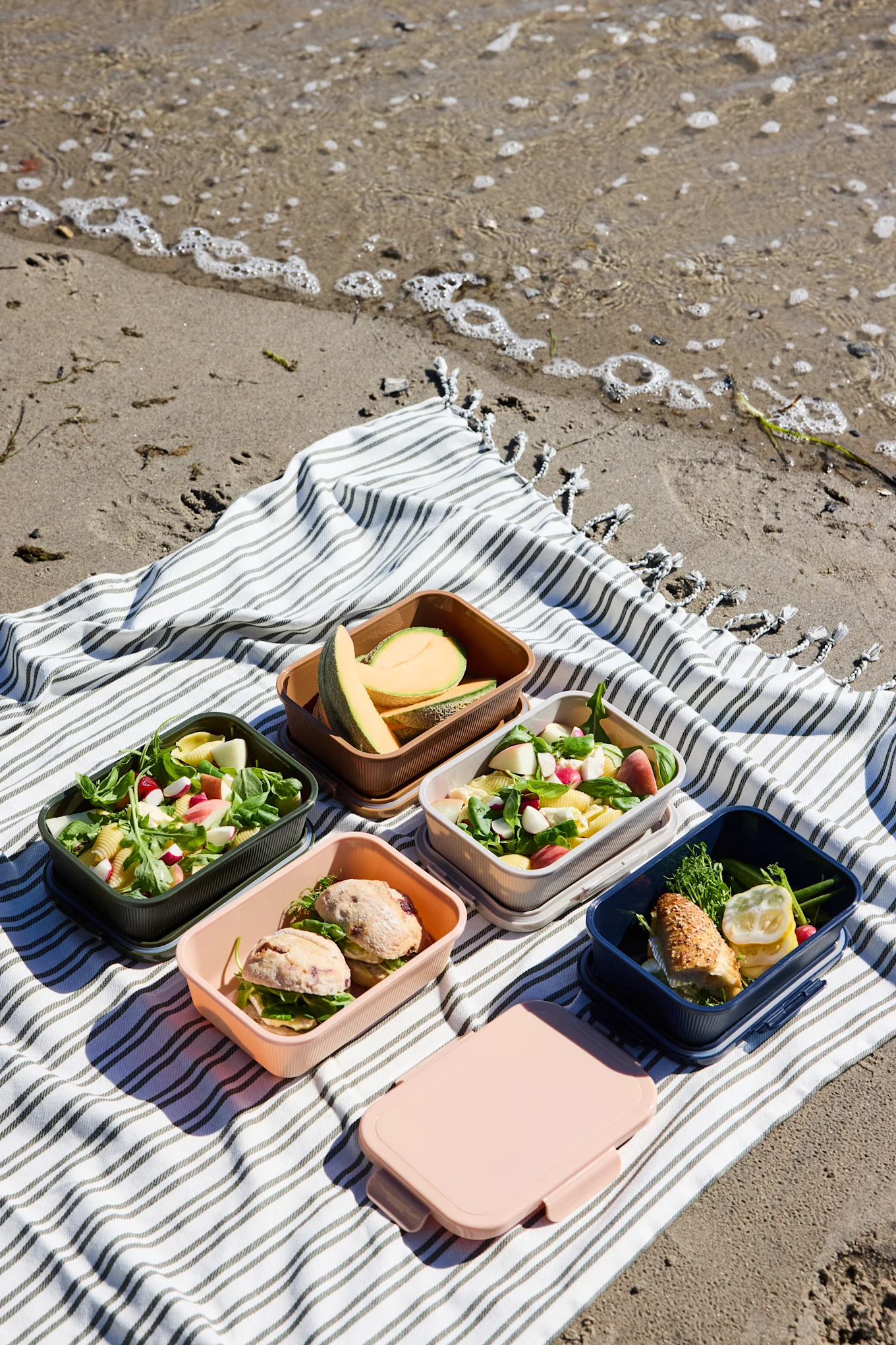 Pięć otwartych, kolorowych pojemników na lunch z sałatkami, kanapkami i melonem, plus jedno zamknięte różowe pudełko, na pasiastym kocu na plaży.