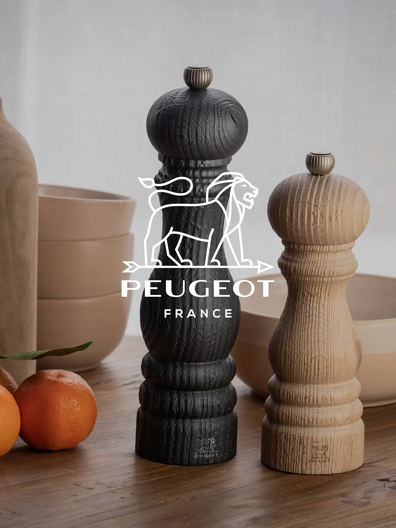 Dwa młynki do pieprzu Peugeot, jeden czarny i jeden z naturalnego drewna, na drewnianym stole z mandarynkami i miskami.