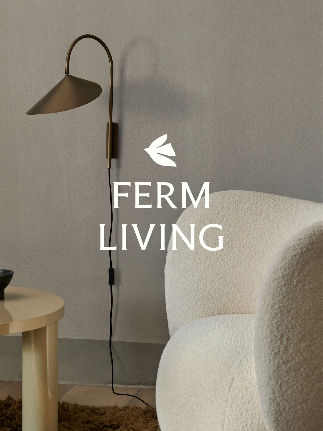 Logo Ferm Living nad mosiężną lampą ścienną, kremowym fotelem bouclé i jasnym drewnianym stolikiem bocznym w minimalistycznym wnętrzu.