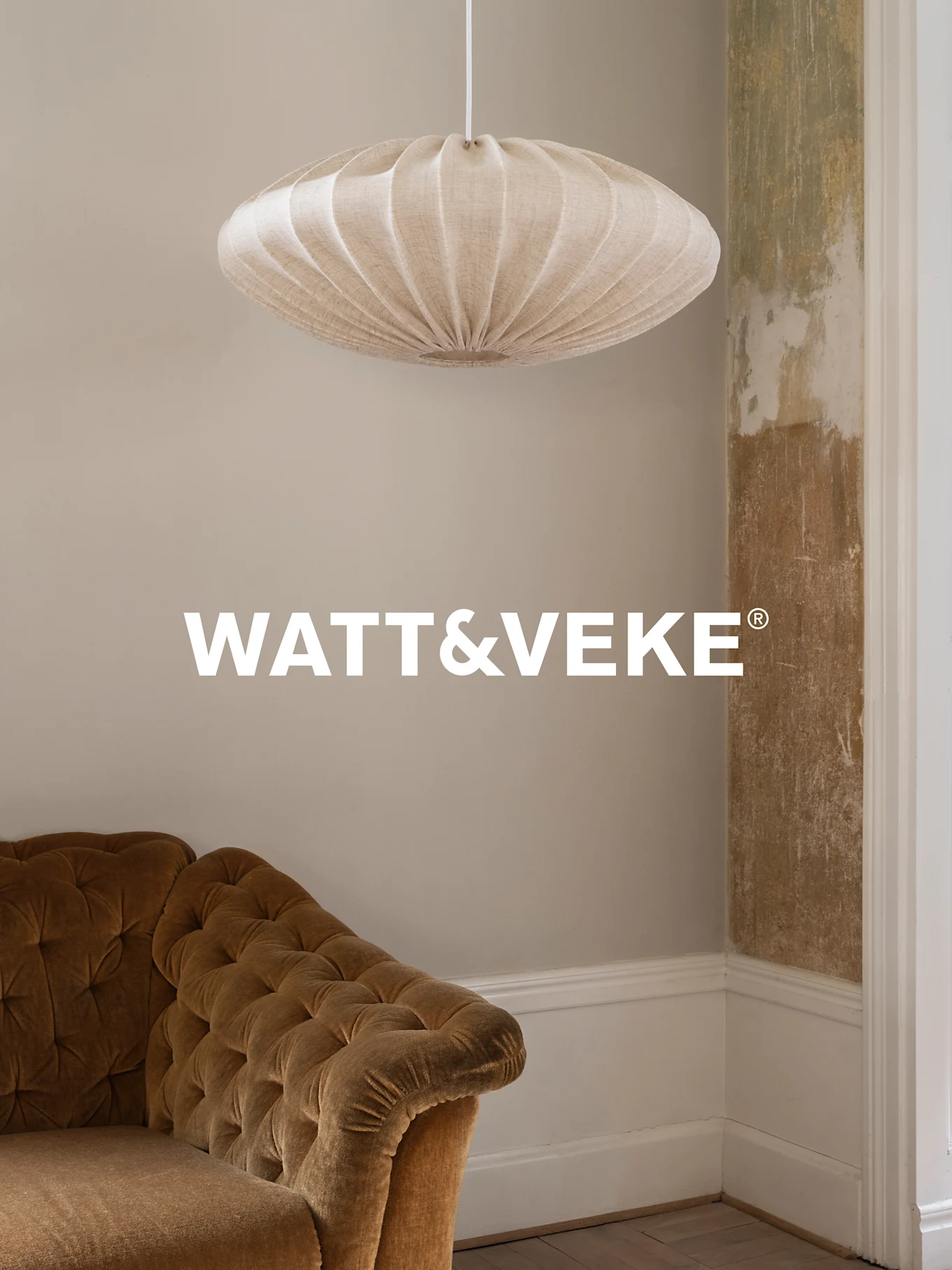 Beżowa plisowana lampa wisząca wisi nad brązową aksamitną sofą w pokoju ze zniszczoną ścianą.