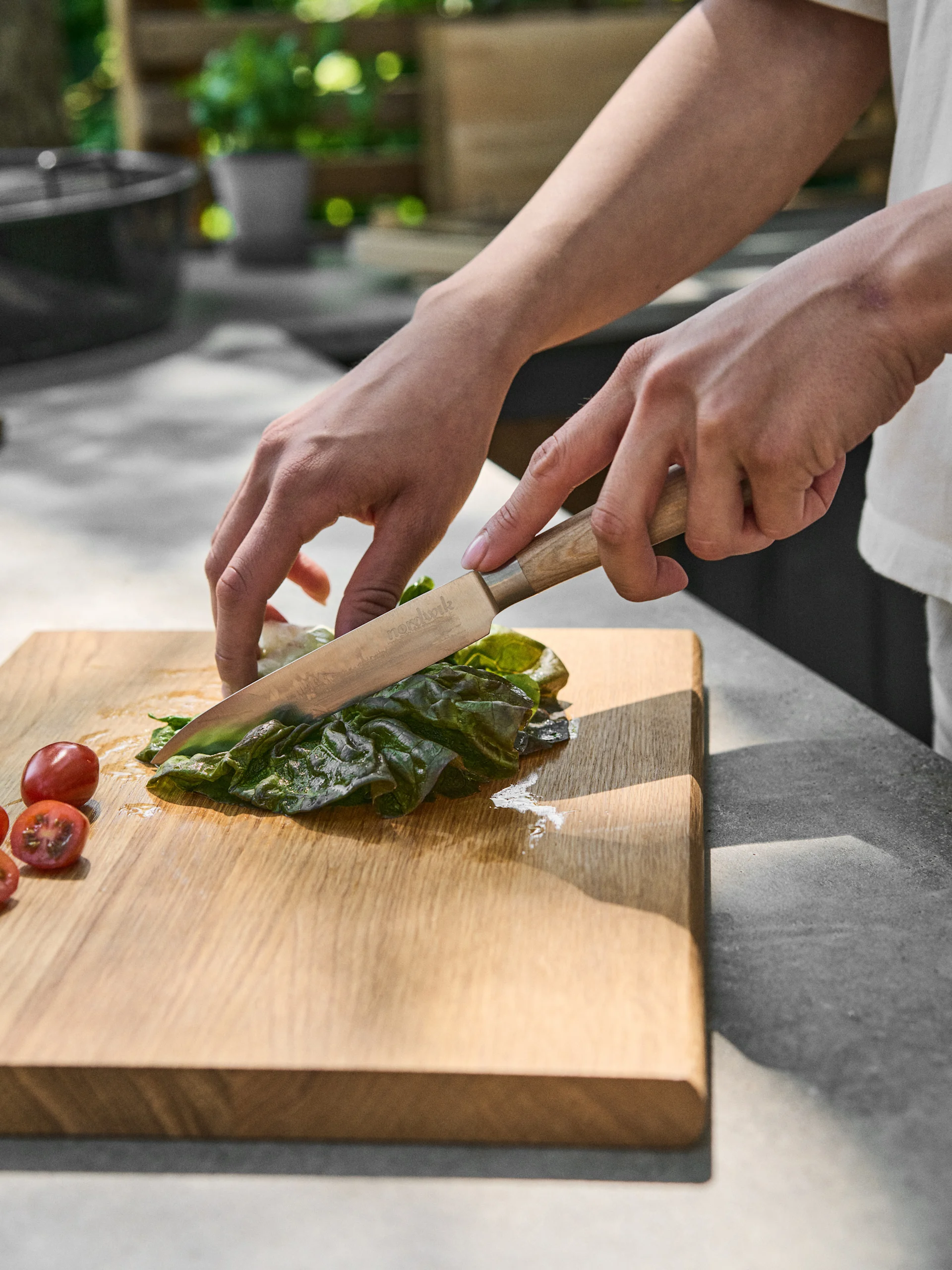 Ręce kroją zielony liść nożem na drewnianej desce, obok pomidorków koktajlowych.