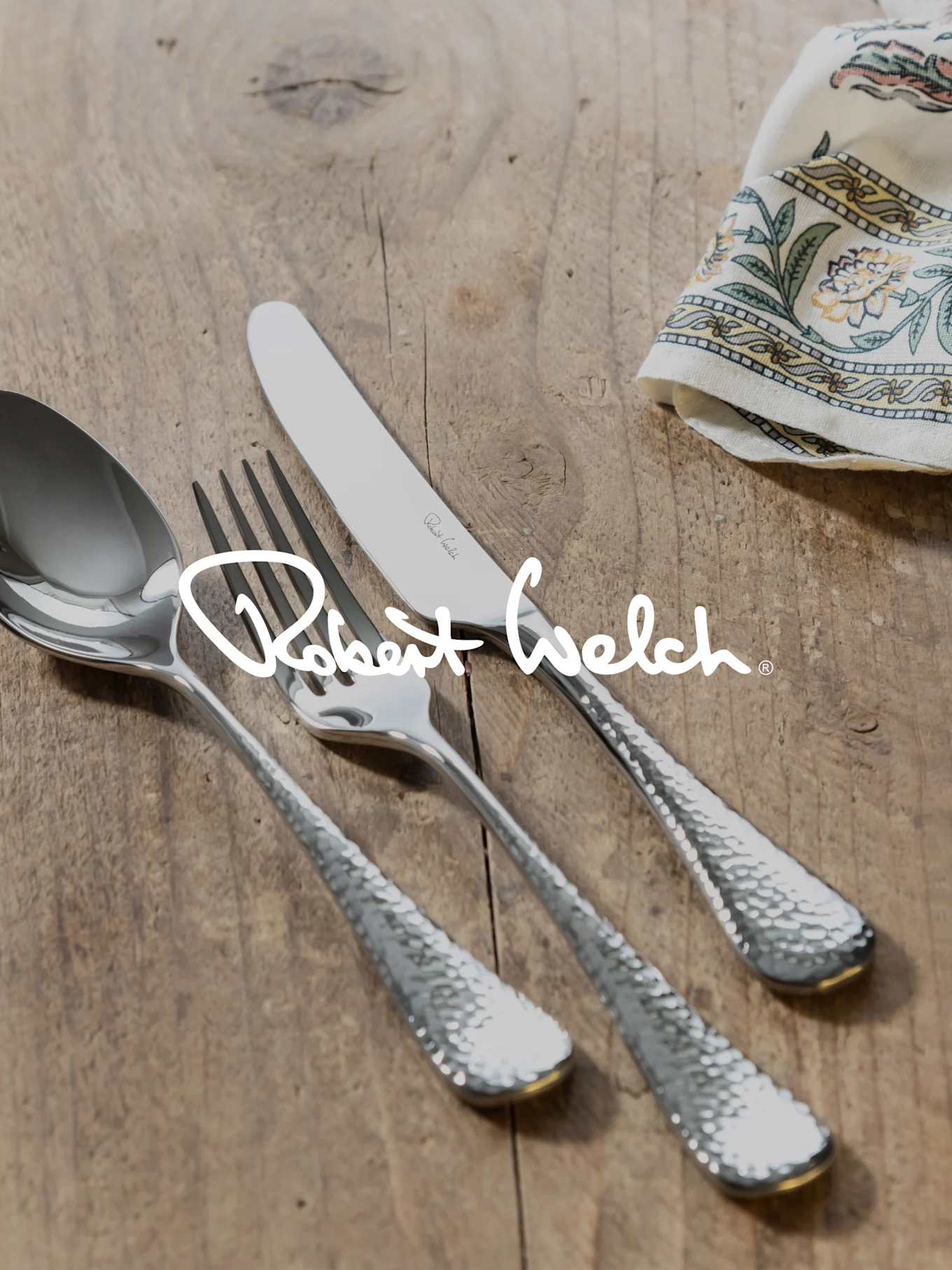 Logo Robert Welch na srebrnych młotkowanych sztućcach (łyżka, widelec, nóż) na drewnie, wzorzysta ściereczka.