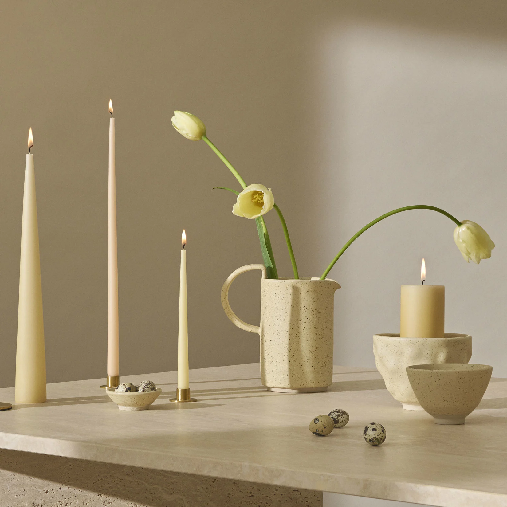 Martwa natura z zapalonymi świecami, jasnożółtymi tulipanami w nakrapianym dzbanku i przepiórczymi jajami na teksturowanej powierzchni.