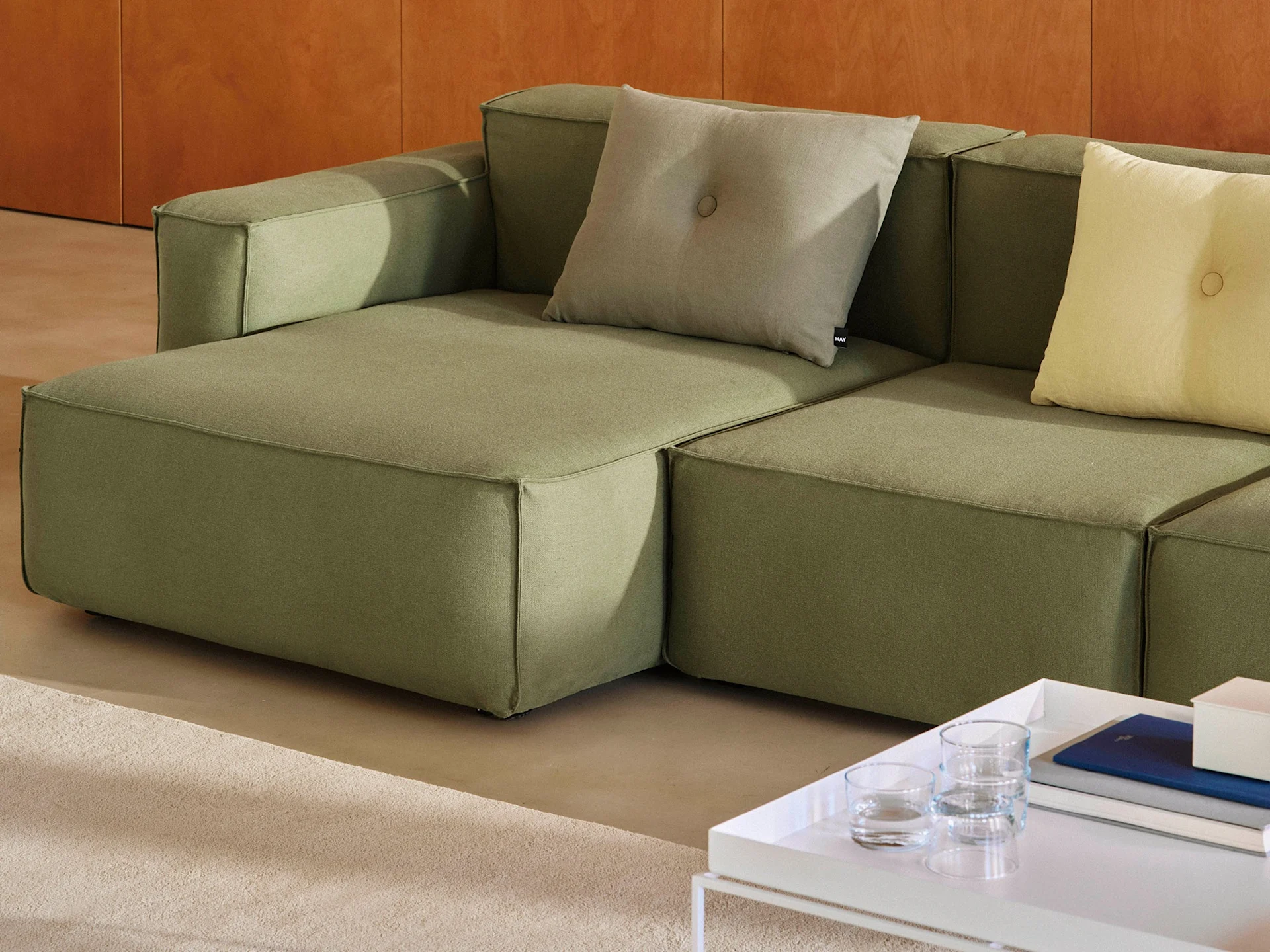 Modułowa oliwkowozielona sofa z jasno-szarymi i żółtymi poduszkami dekoracyjnymi, obok białego stolika kawowego z książkami.