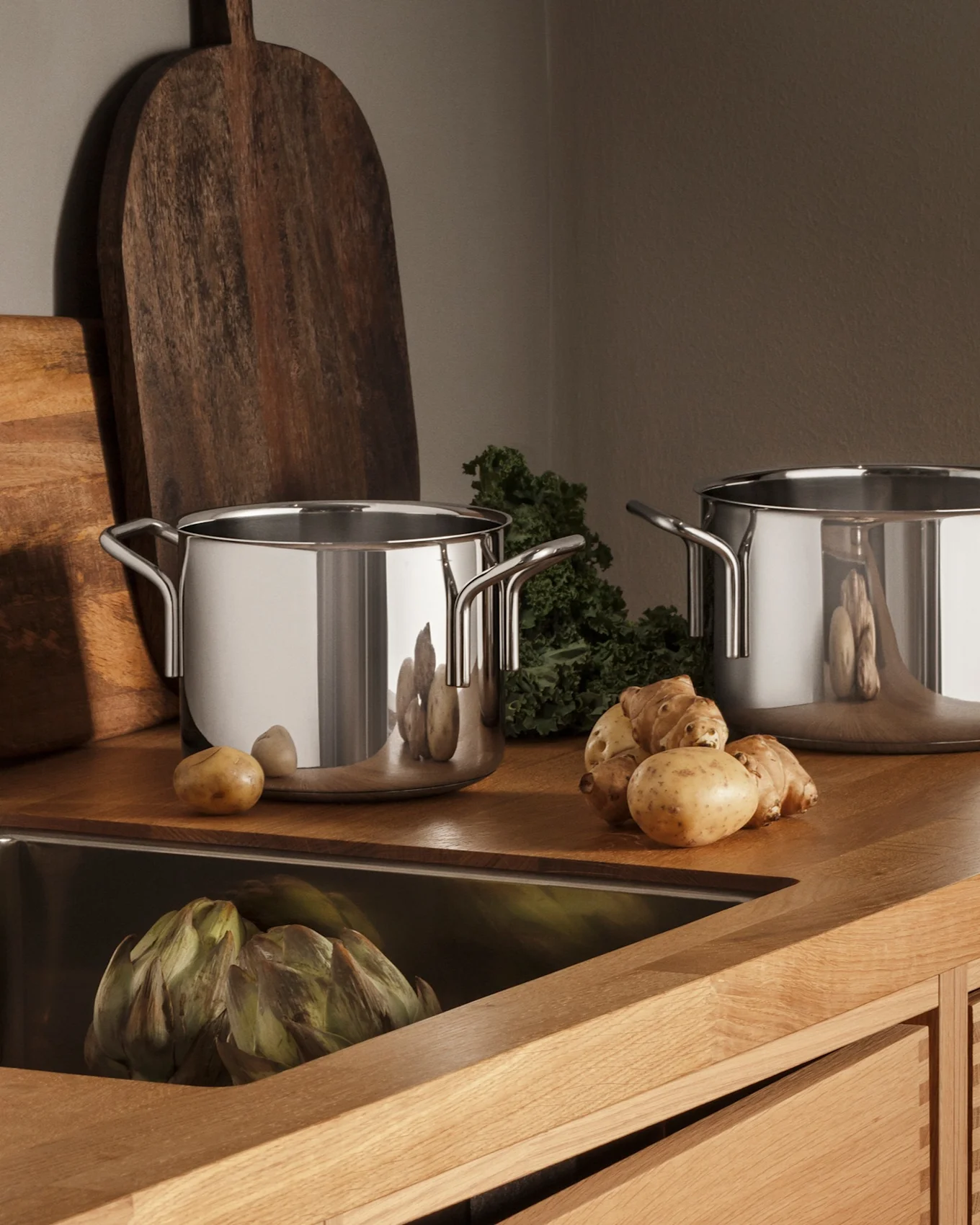 Nowoczesna kuchnia z drewnianym blatem, zlewozmywakiem ze stali nierdzewnej, kranem, trzema garnkami i świeżymi produktami, takimi jak ziemniaki i karczochy.