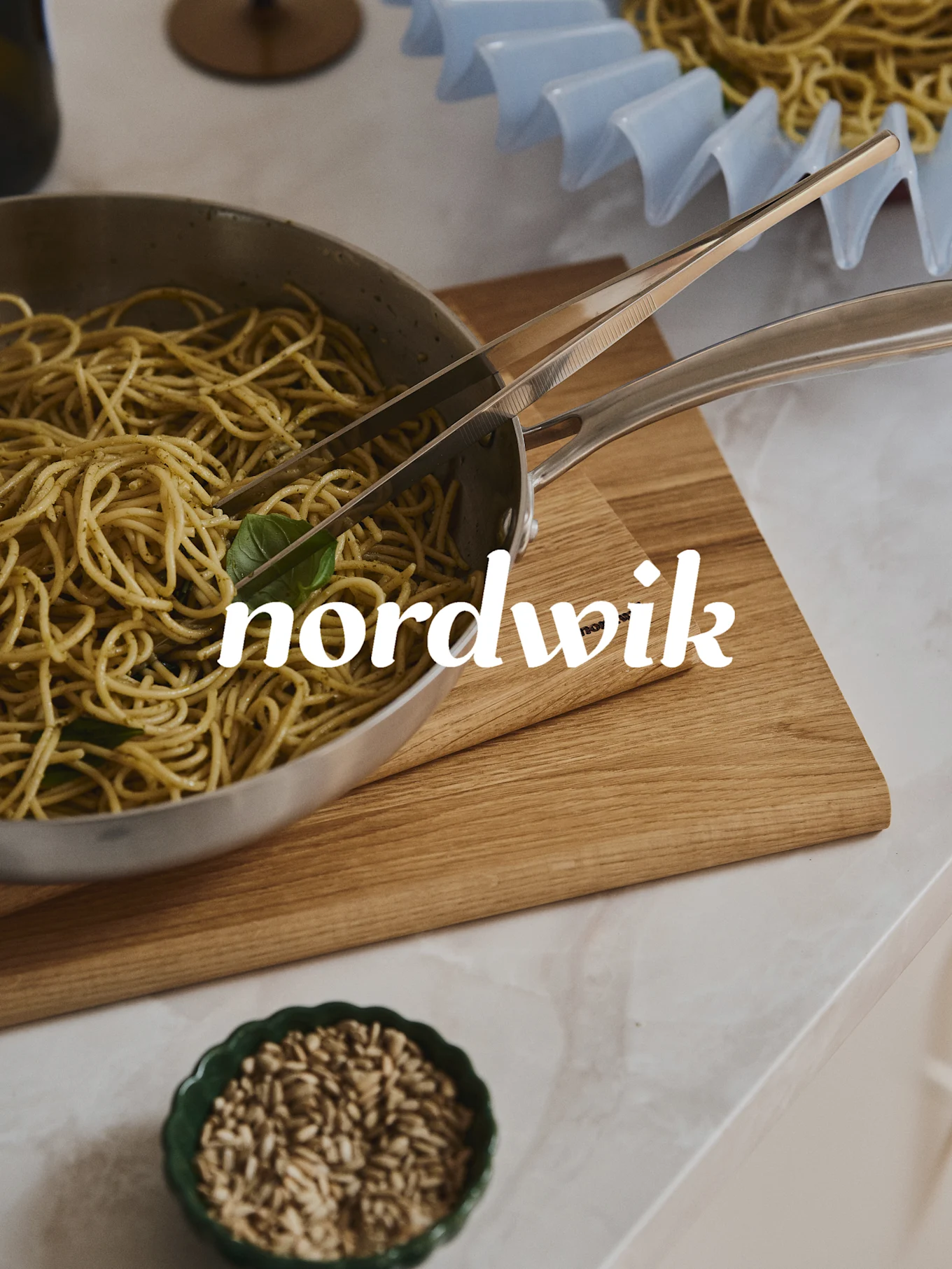 Spaghetti z pesto w stalowej patelni z szczypcami na drewnianej desce. Poniżej miska nasion, a w tle suszarka do makaronu. Nakładka tekstowa 'nordwik'.
