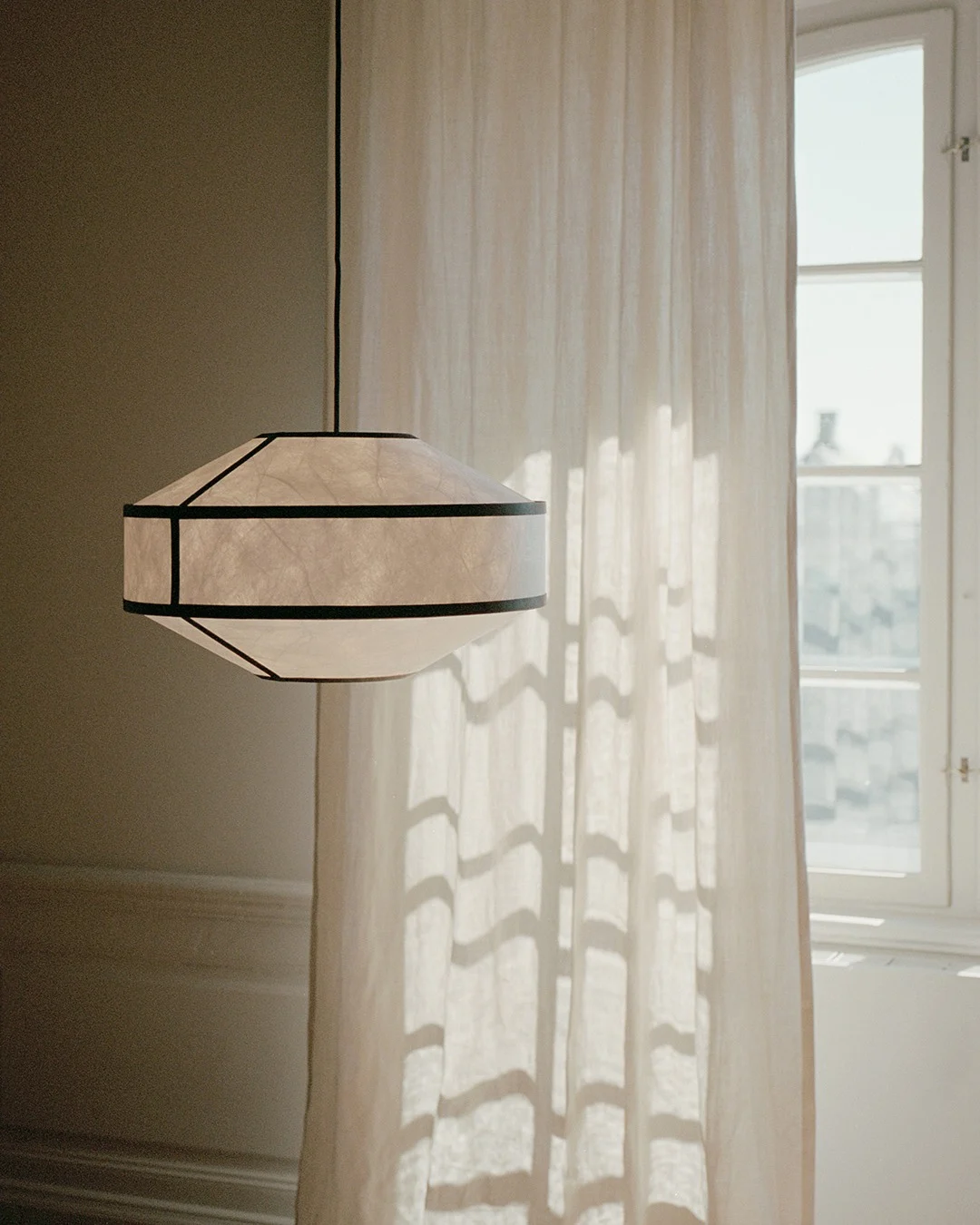 Geometryczna papierowa lampa wisząca z ciemnymi ramkami wisi obok zasłony i okna oświetlonych słońcem.