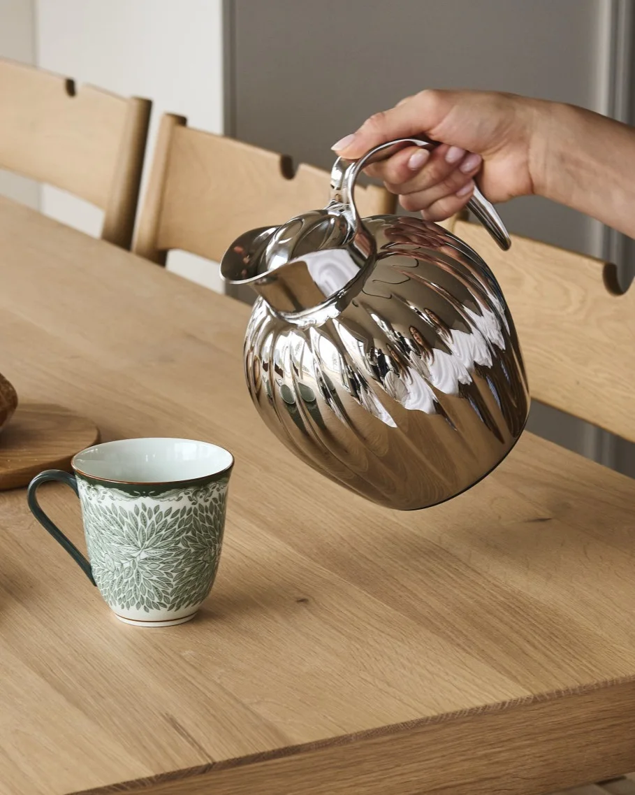 Dłoń nalewa ze srebrnej karbowanej karafki do białego kubka z zielonym wzorem liści na drewnianym stole.