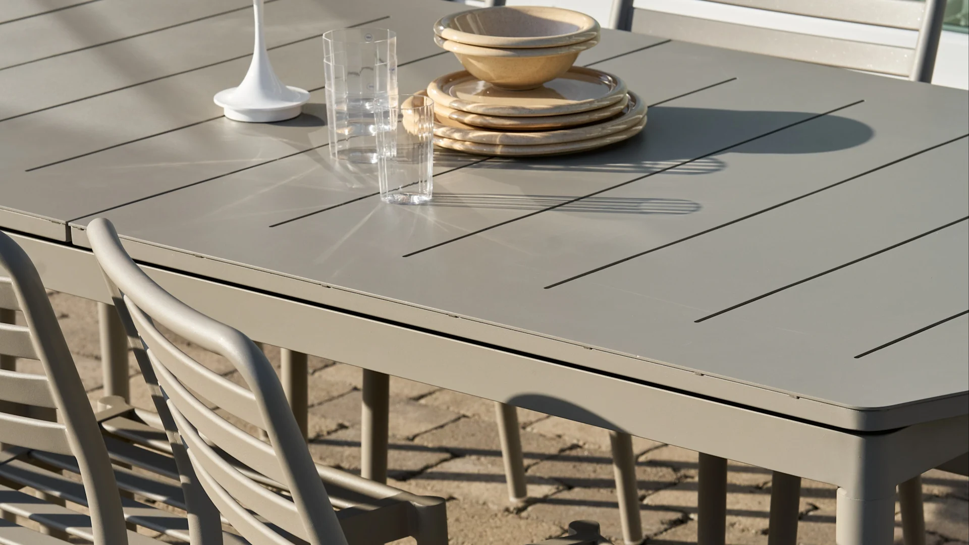 Jasnoszary lamelowy stół obiadowy na zewnątrz ze stosami beżowych talerzy i misek, dwoma przezroczystymi szklankami i białą ozdobą.
