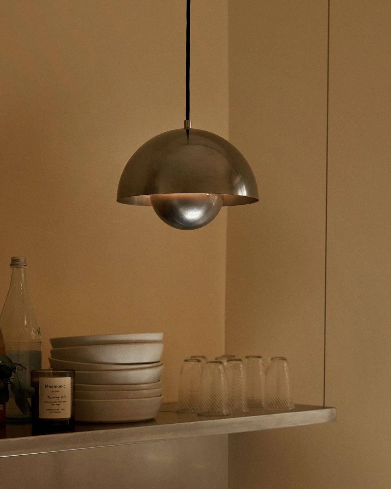 Metalowa lampa wisząca w kształcie kopuły wisi nad półką z miskami, szklankami, butelkami, owocem i świecą.