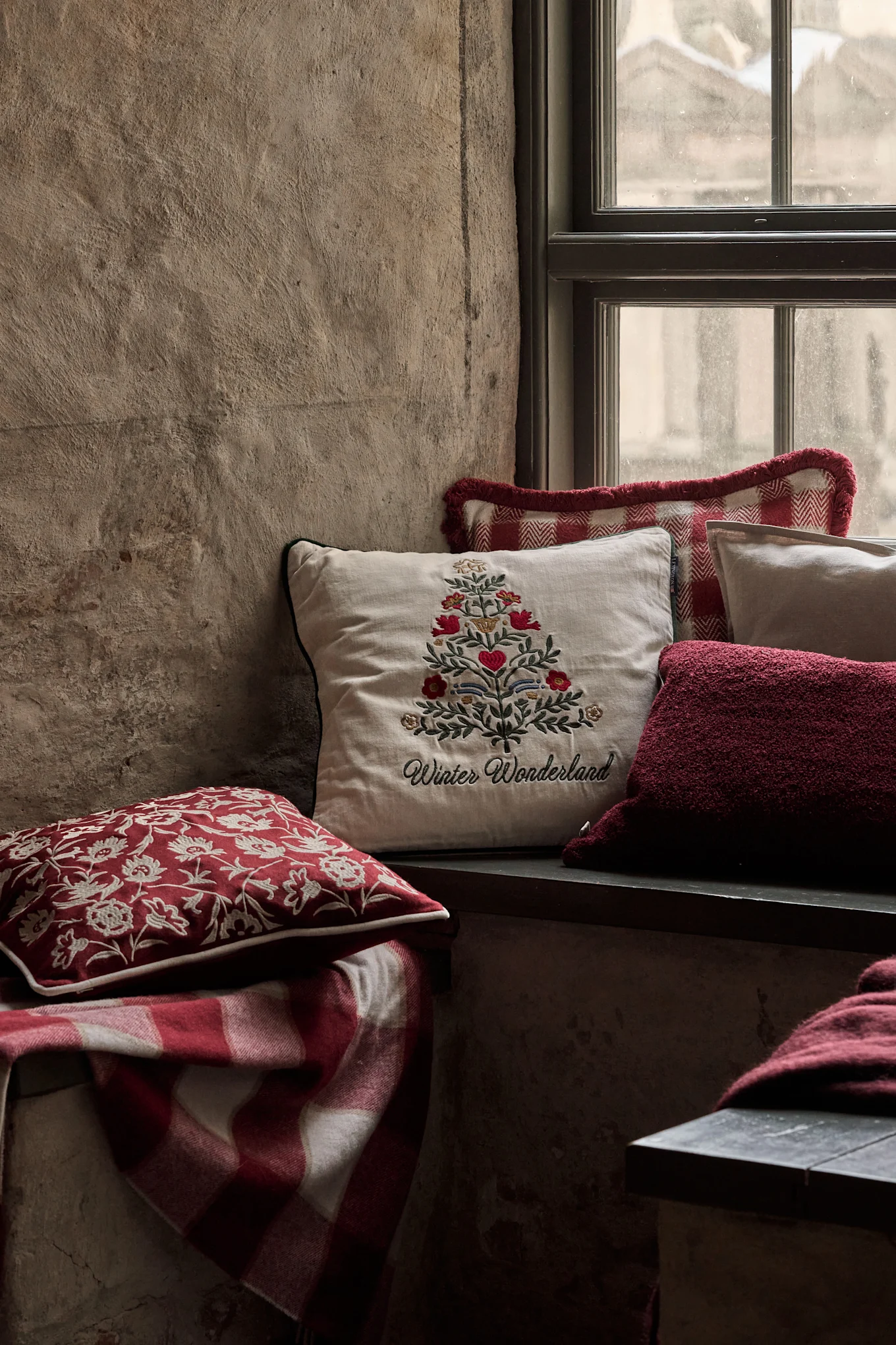 Przytulny kącik okienny z świątecznymi poduszkami, w tym jedną z haftowaną choinką i napisem 'Winter Wonderland', oraz czerwonym kocem w kratę.
