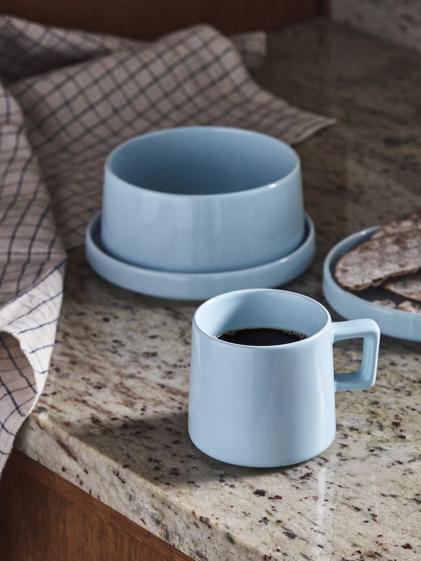 Jasnoniebieski ceramiczny kubek kawy, miska i talerz z podpłomykami na granitowym blacie.