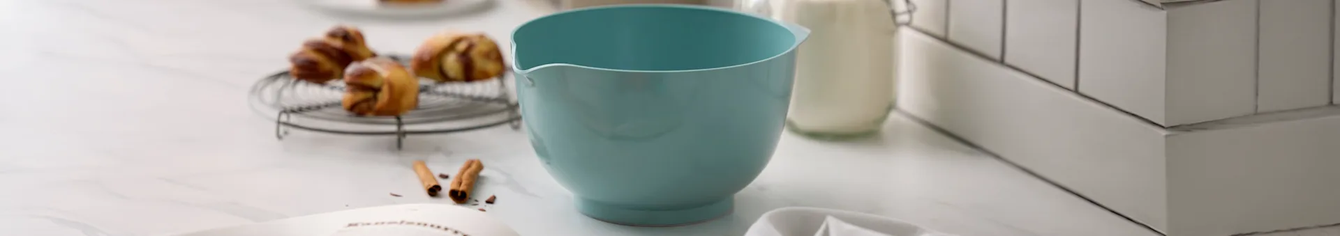 Jasnoniebieska miska do mieszania, wypieki na kratce, butelka mleka i książka kucharska na marmurowym blacie.