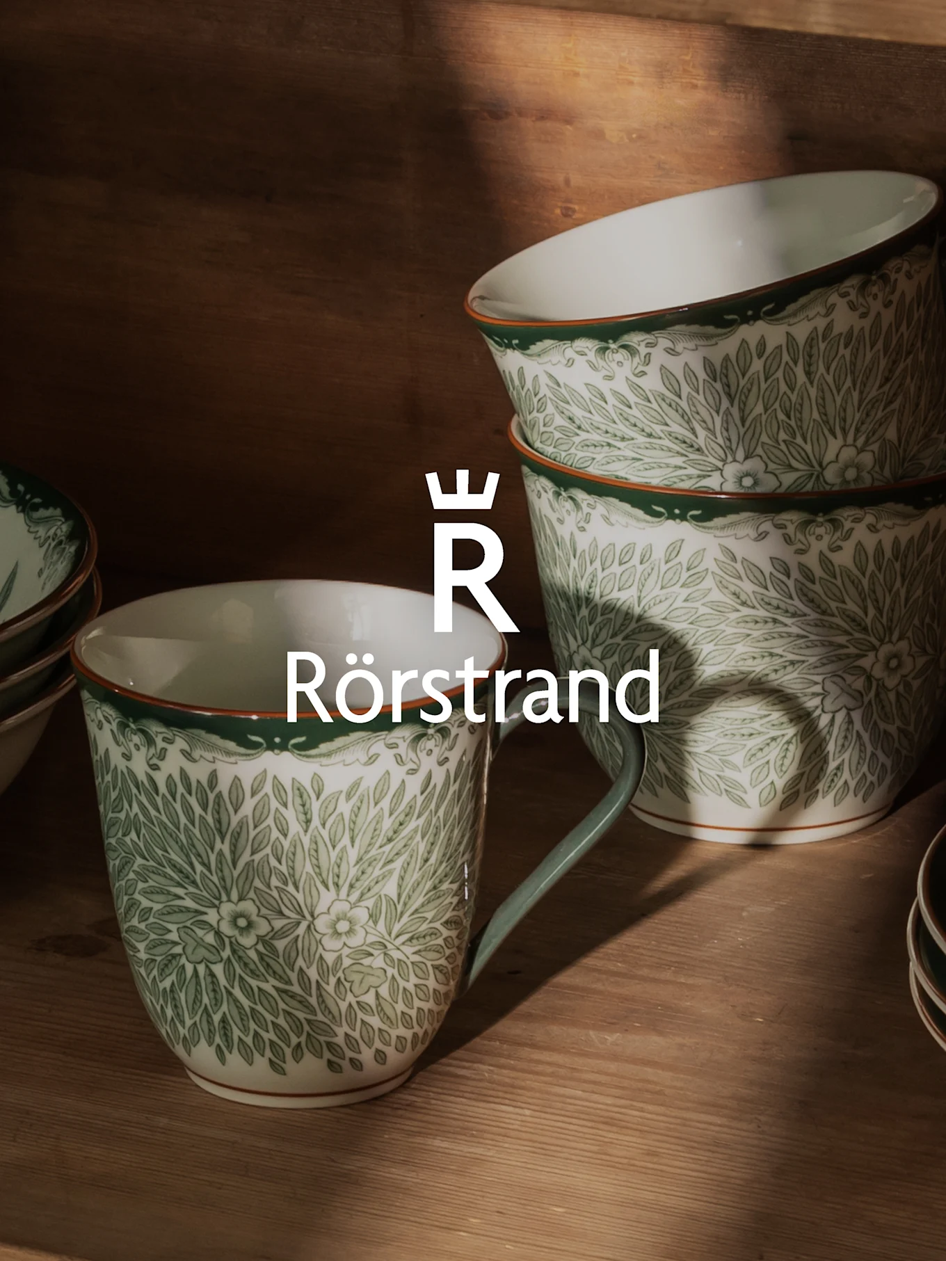 Białe ceramiczne kubki i miski z zielonym wzorem liści, pomarańczową obwódką i logo Rörstrand.